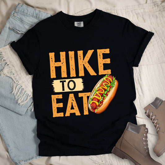 Black t-shirt with "HIKE TO EAT" in distressed orange lettering. The word "TO" appears on a beige background between "HIKE" and "EAT". A colorful hot dog illustration with mustard, ketchup and toppings appears next to the "T" in "EAT". Features a classic crew neck and casual rolled sleeves, styled alongside light wash denim and brown hiking boots. 