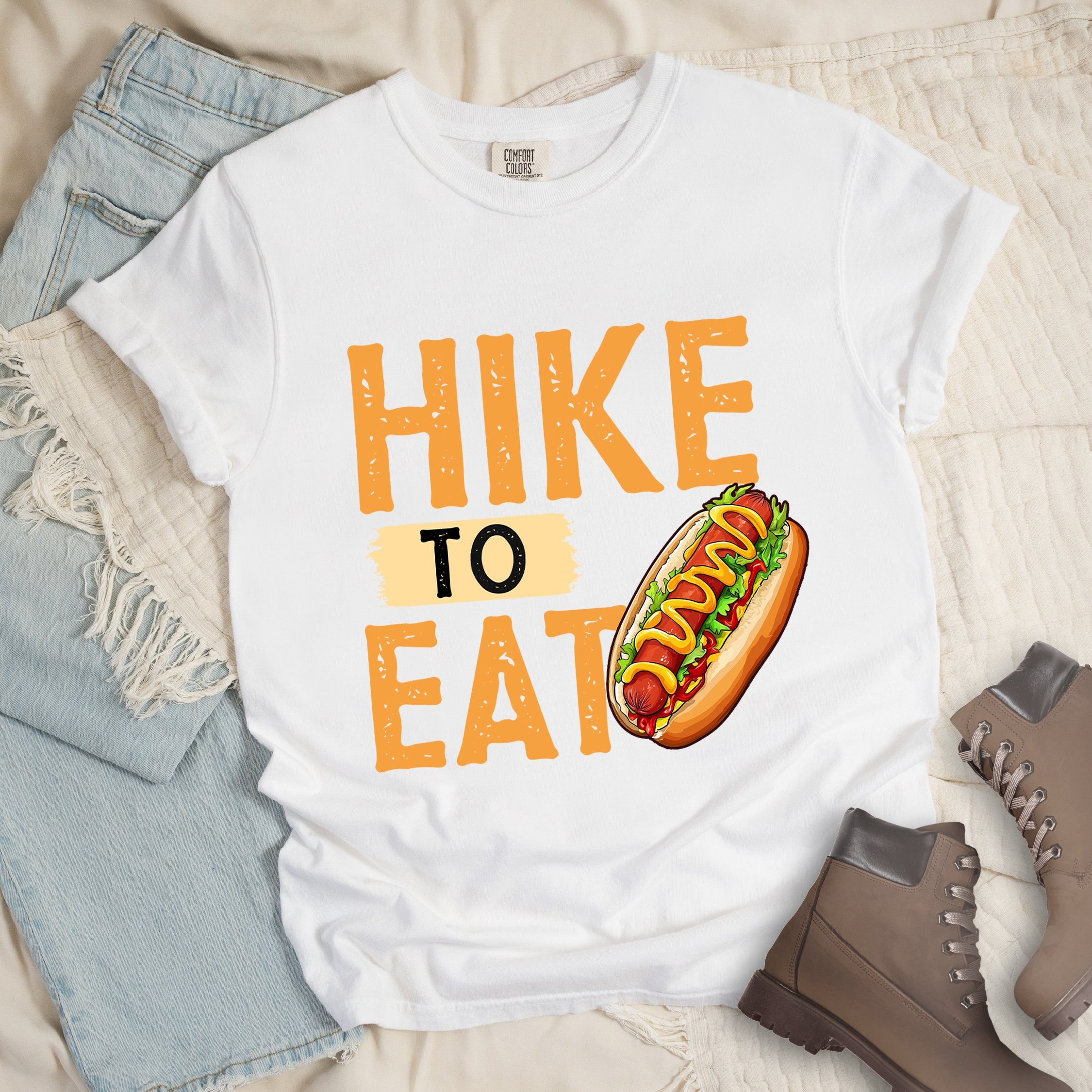White shirt showing a hiking design with 'HIKE TO EAT phrase and an illustration of a hotdog in a bun with mustard on top