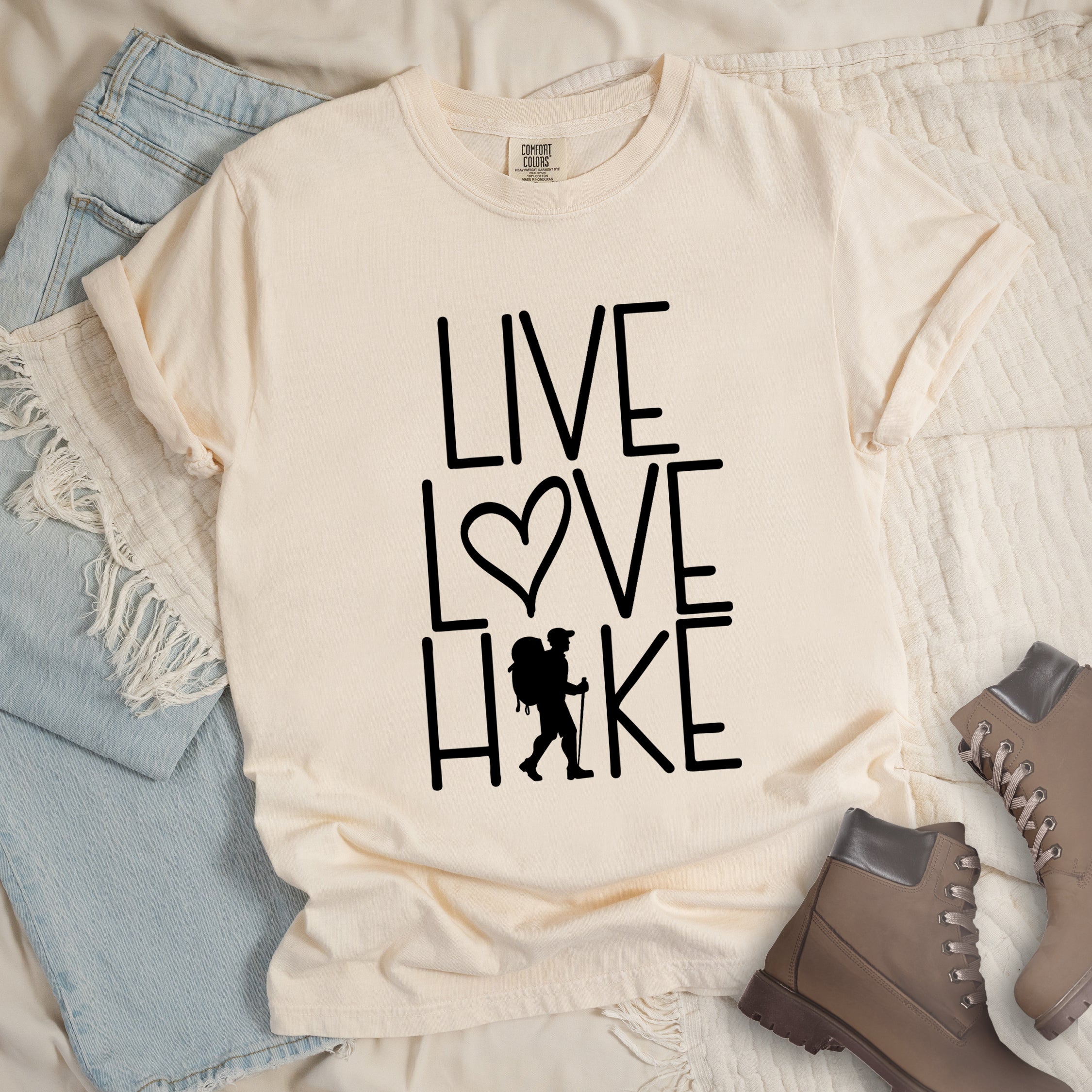 Ivory (cream) colored t-shirt with black text reading 'Live Love Hike' featuring a heart symbol and a male hiker silhouette with backpack, styled with light wash denim jeans and brown hiking boots on white bedding. 