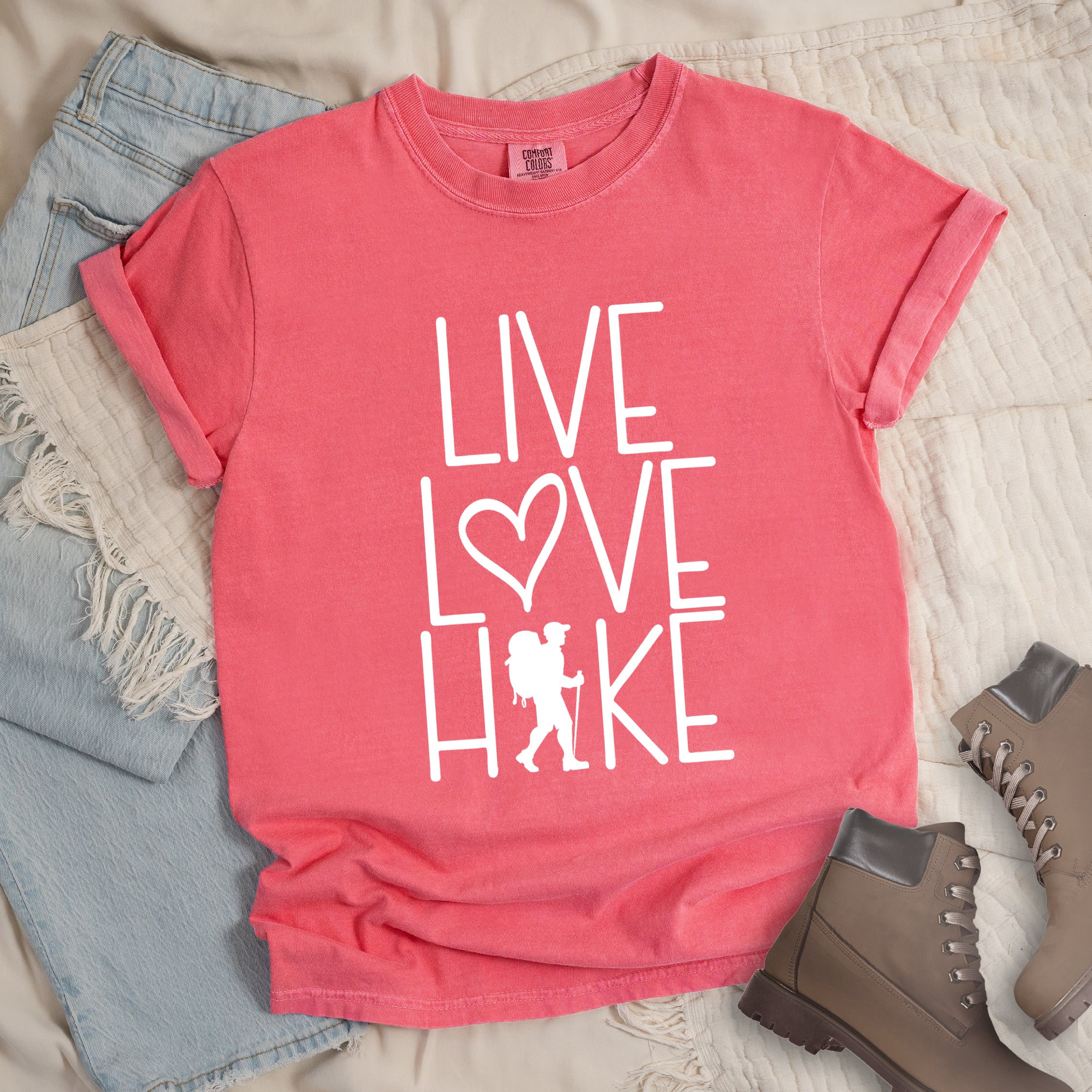 Watermelon colored t-shirt with white text reading 'Live Love Hike' featuring a heart symbol and a male hiker silhouette with backpack, styled with light wash denim jeans and brown hiking boots on white bedding. 
