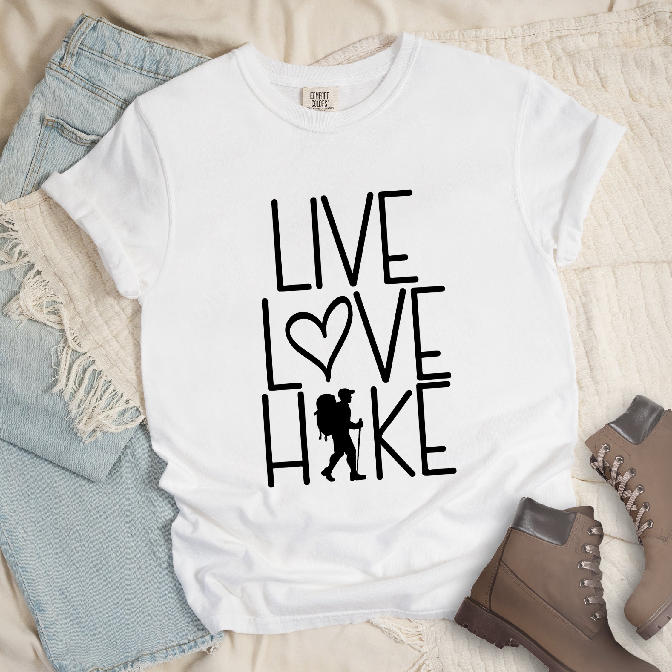 White colored t-shirt with black text reading 'Live Love Hike' featuring a heart symbol and a male hiker silhouette with backpack, styled with light wash denim jeans and brown hiking boots on white bedding. 