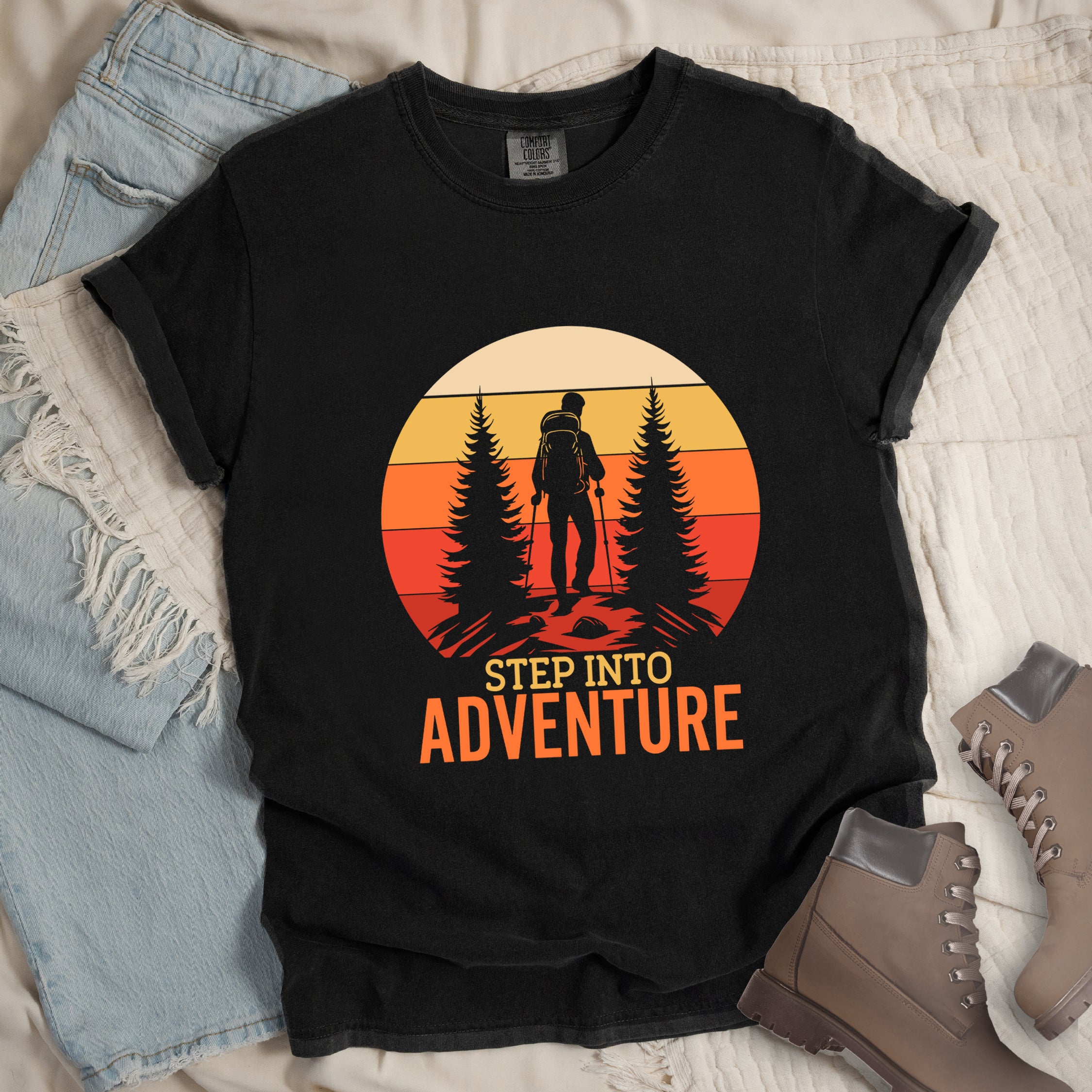 Black graphic t-shirt with 'Step Into Adventure' text and vintage sunset design featuring hiker silhouette among pine trees, styled with light wash jeans and brown hiking boots
