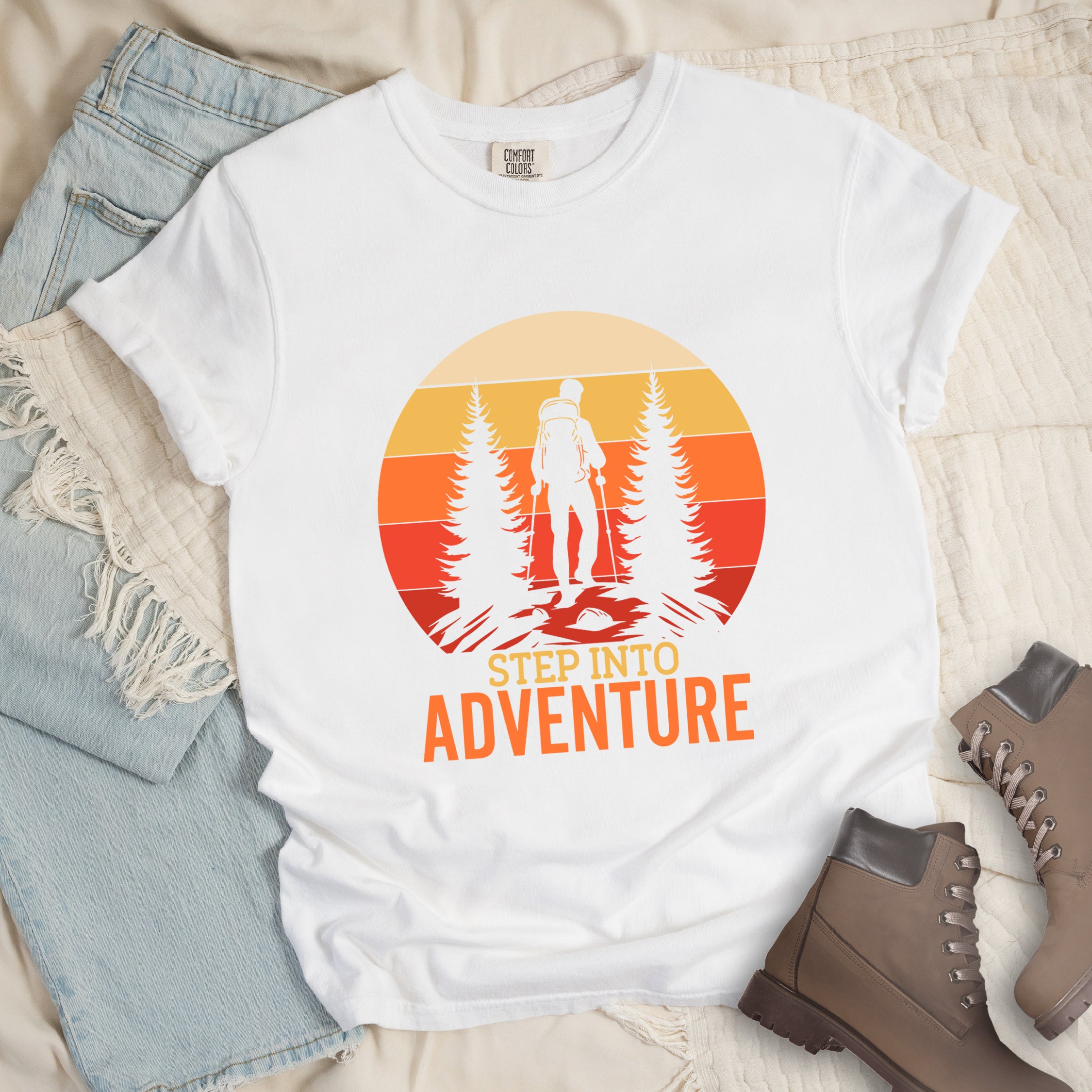 White graphic t-shirt with 'Step Into Adventure' text and vintage sunset design featuring hiker silhouette among pine trees, styled with light wash jeans and brown hiking boots