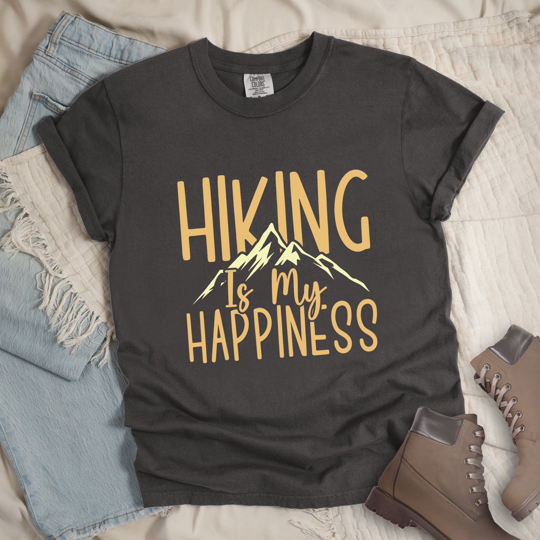 Pepper (Dark Gray) graphic t-shirt with 'Hiking Is My Happiness' text in yellow and cream mountain design, styled with light wash jeans and brown hiking boots