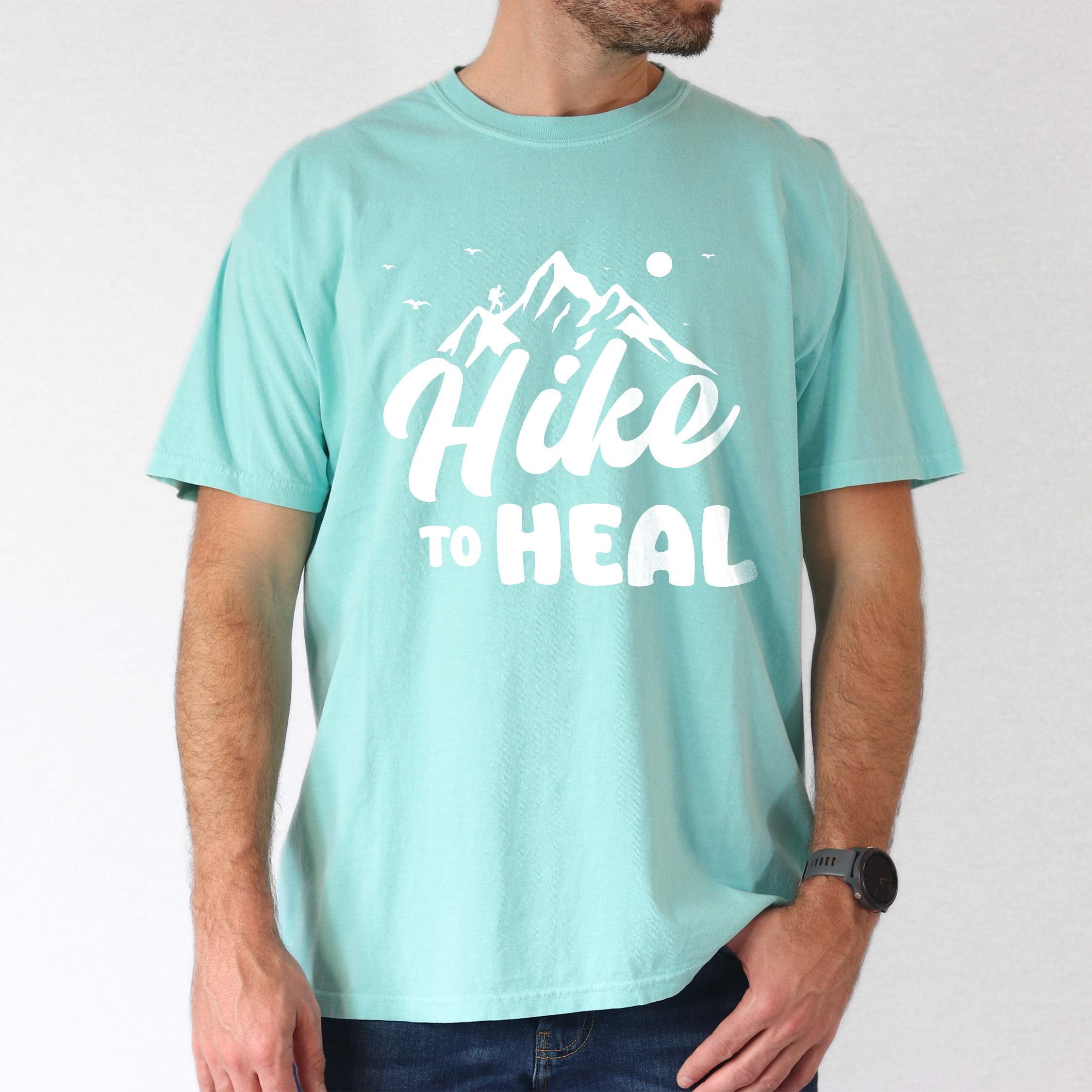 Man wearing chalky mint "Hike to Heal" colored statement tee with bold typography, mountain, sunrise, perfect for adventure driven individuals.