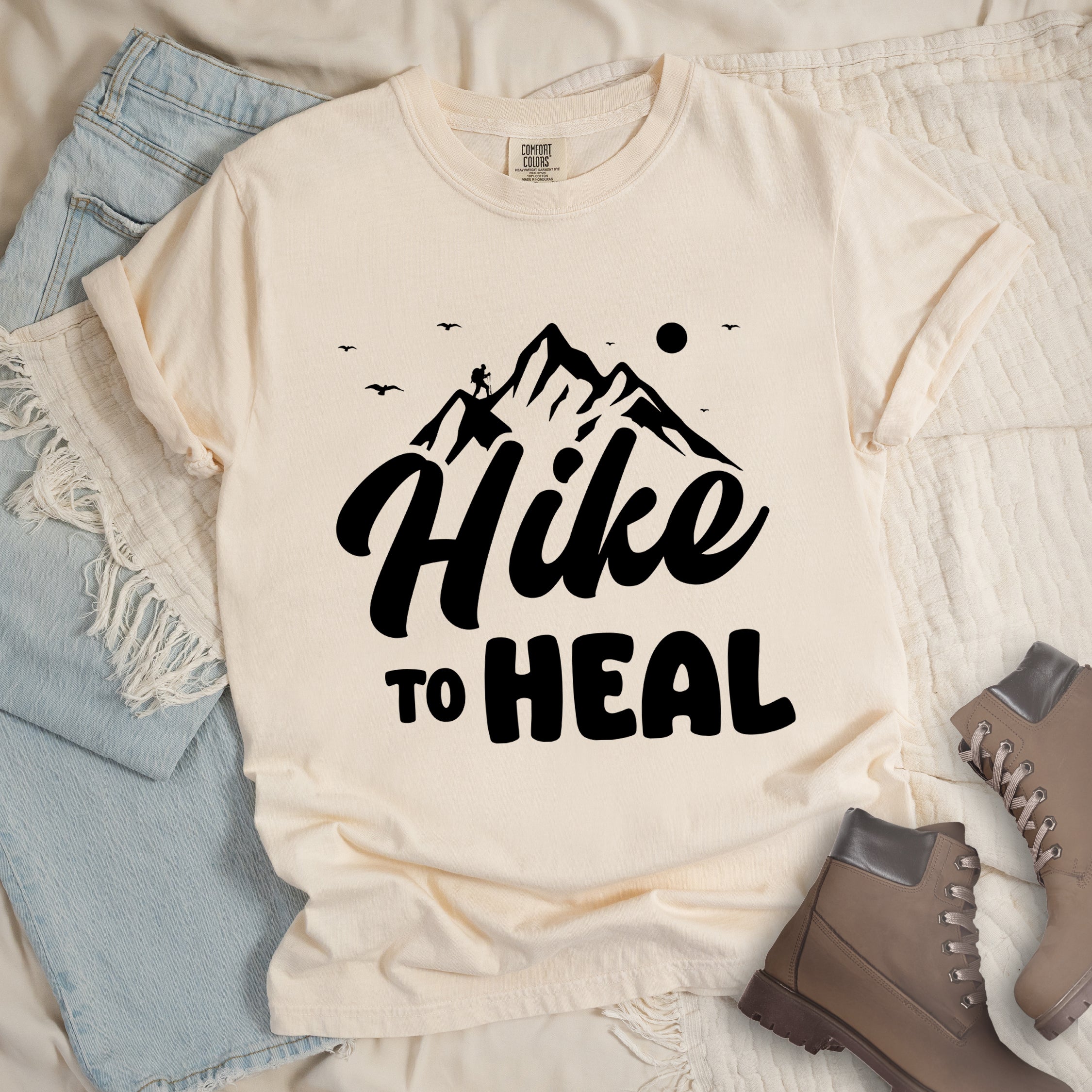 Ivory (Cream White) graphic t-shirt with 'Hike to Heal' text in black with mountain and bird design, styled with light wash jeans and brown hiking boots