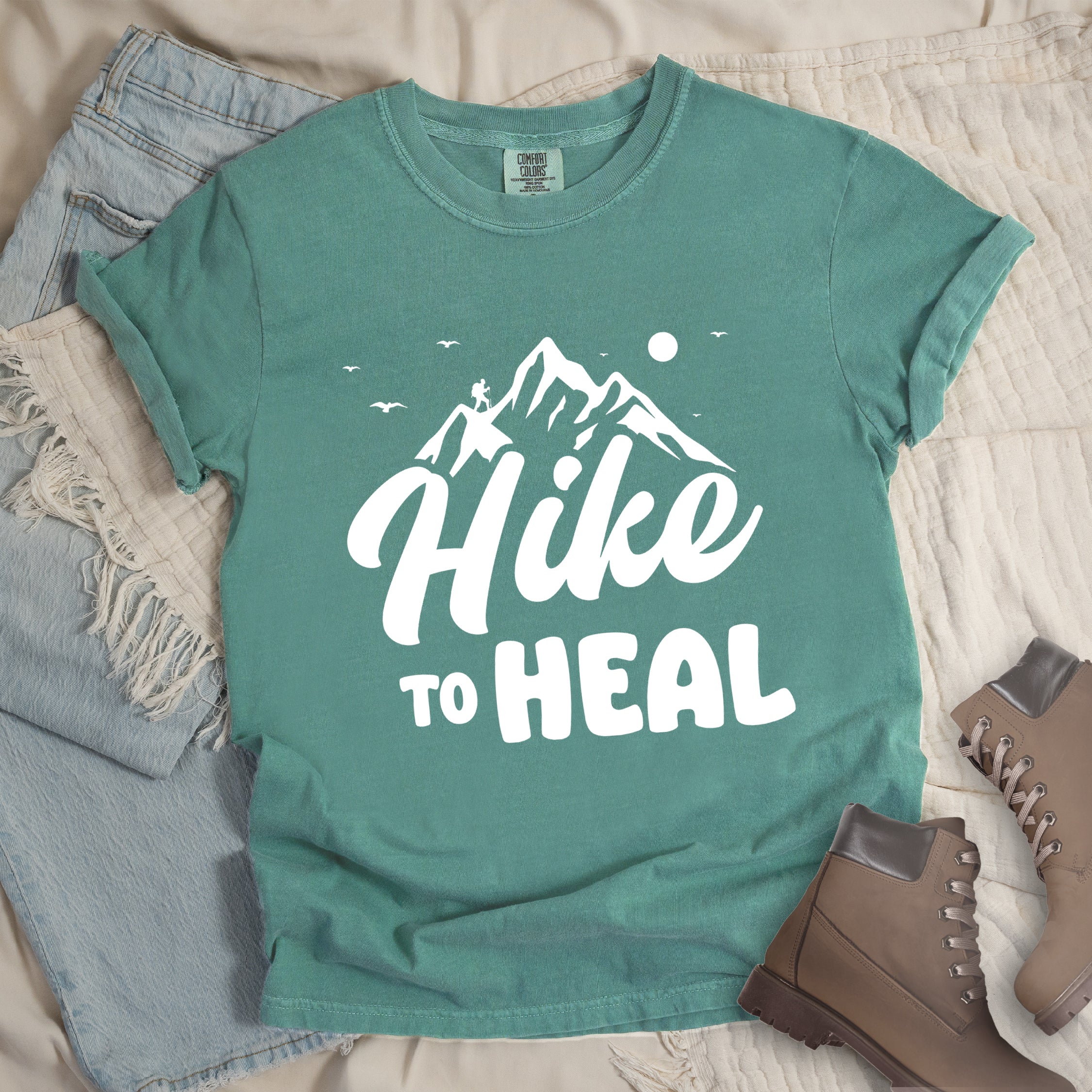 Light green graphic t-shirt with 'Hike to Heal' text in white with mountain and bird design, styled with light wash jeans and brown hiking boots