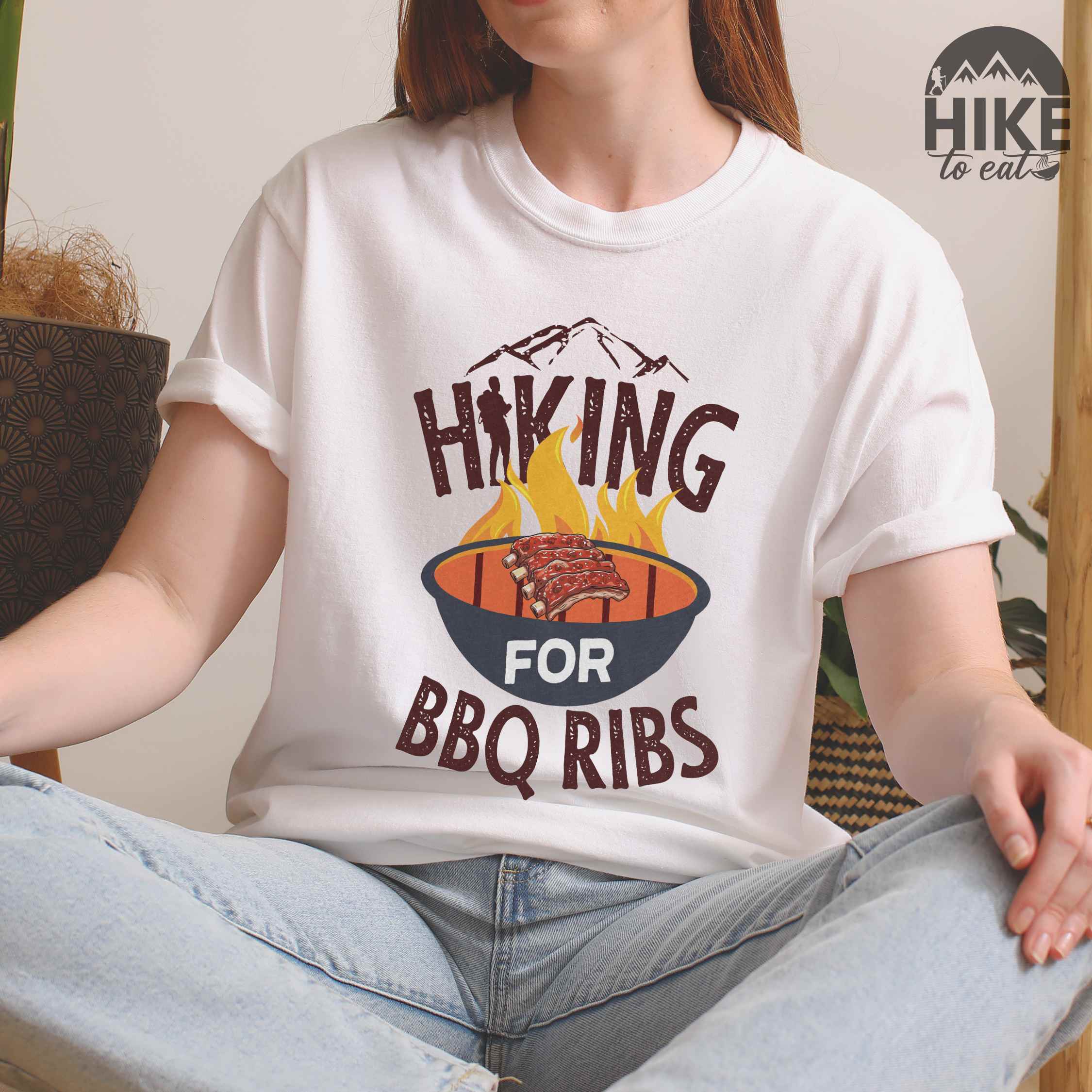 Woman wearing white Hiking for BBQ ribs colored vintage tee with bold typography, illustrated grilling bbq ribs, mountains, birds, and bbq enthusiasts