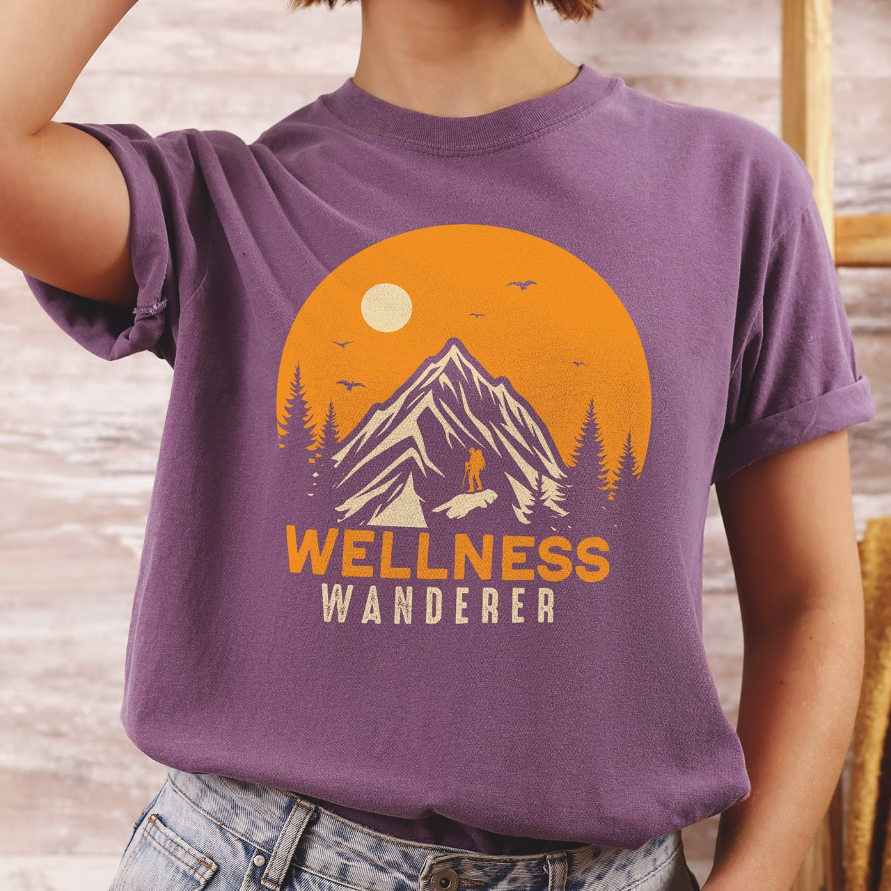 Woman wearing berry (purple) Wellness lifestyle vintage tee featuring mountain peak landscape with character silhouette at summit orange sunset circle flying birds pine trees and bold Wellness Wanderer typography on Comfort Colors shirt for nature lovers seeking balance