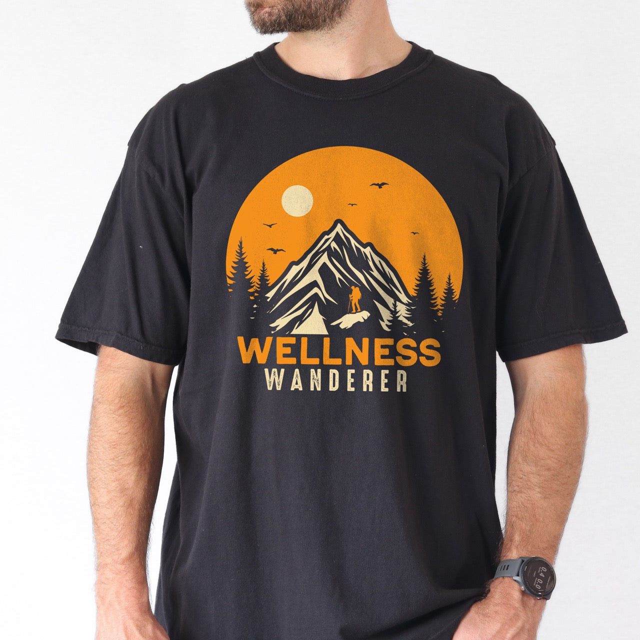 Man wearing black Wellness lifestyle vintage tee featuring mountain peak landscape with character silhouette at summit orange sunset circle flying birds pine trees and bold Wellness Wanderer typography on Comfort Colors shirt for nature lovers seeking balance