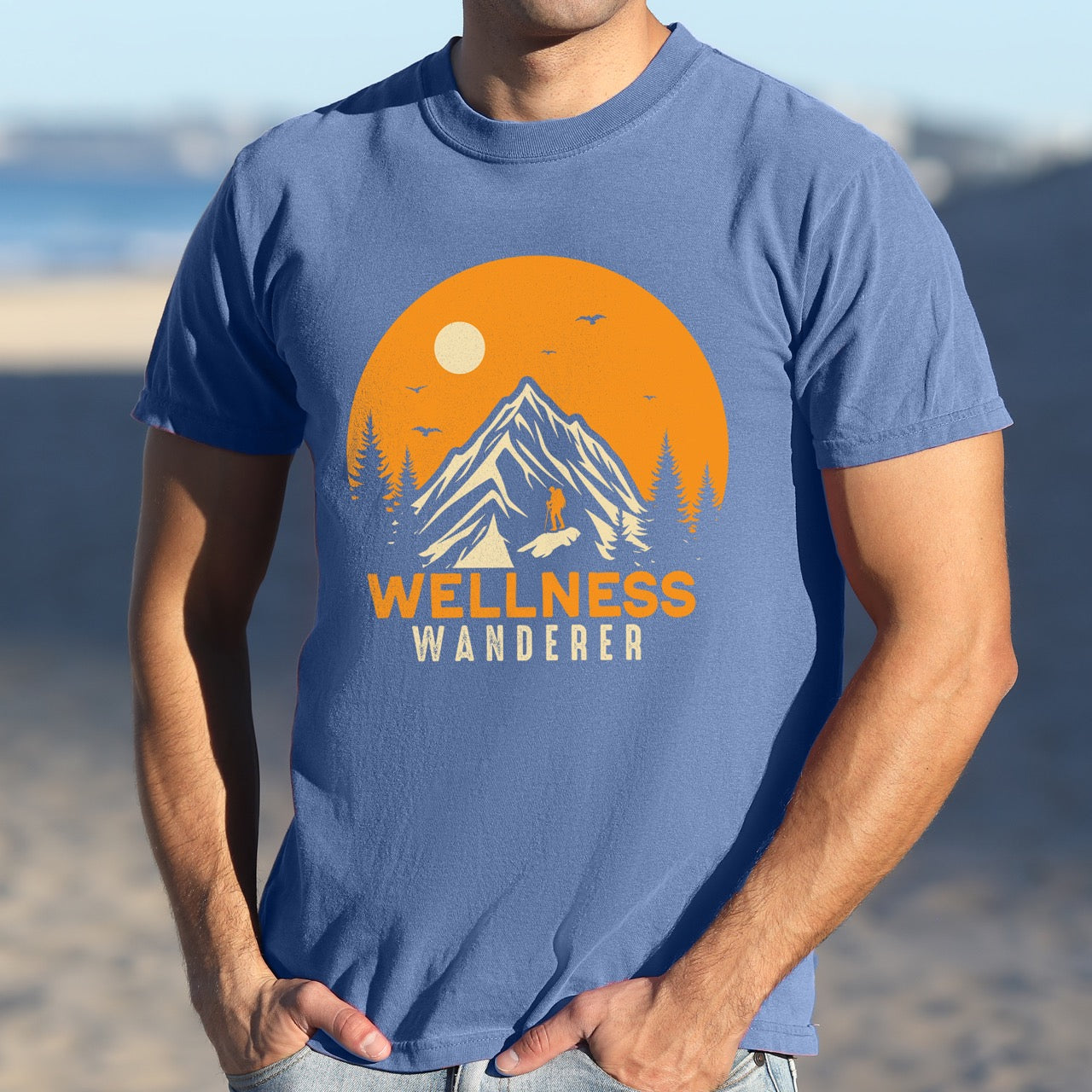 Man wearing blue Wellness lifestyle vintage tee featuring mountain peak landscape with character silhouette at summit orange sunset circle flying birds pine trees and bold Wellness Wanderer typography on Comfort Colors shirt for nature lovers seeking balance