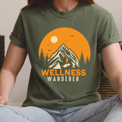 Moss (Dark Green) graphic t-shirt with 'Wellness Wanderer' text in orange lettering featuring circular sunset design with mountains, pine trees, hiker silhouette, flying birds, and sun.