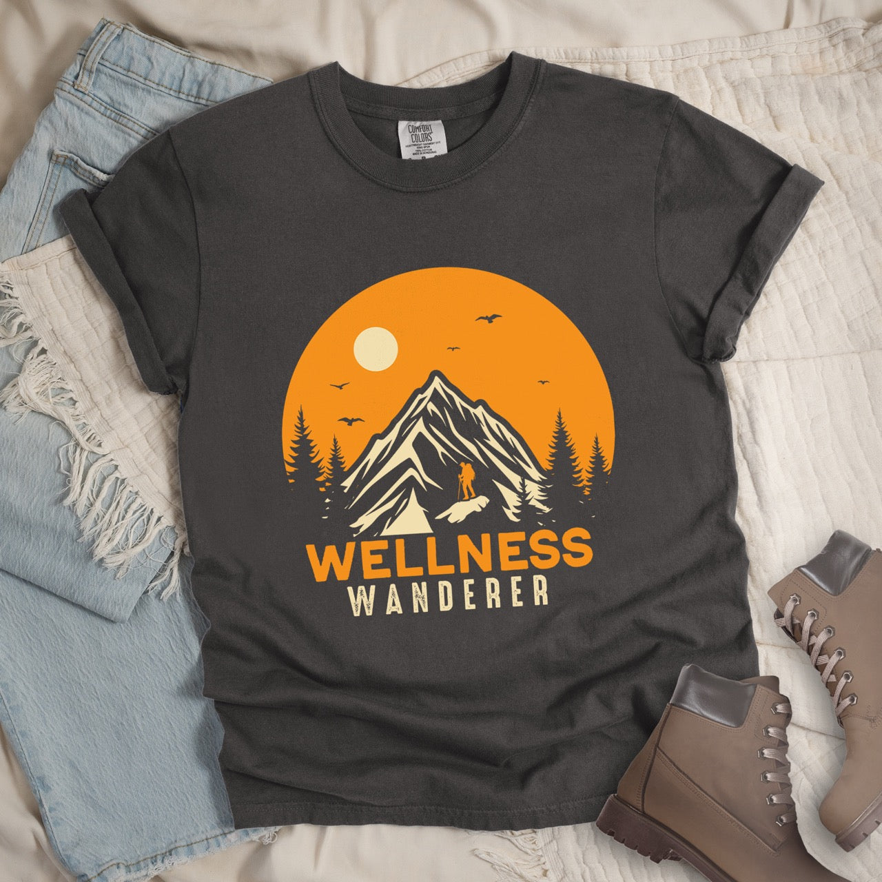 Grey Wellness lifestyle vintage tee featuring mountain peak landscape with character silhouette at summit orange sunset circle flying birds pine trees and bold Wellness Wanderer typography on Comfort Colors shirt for nature lovers seeking balance - laid flat with pants and boots