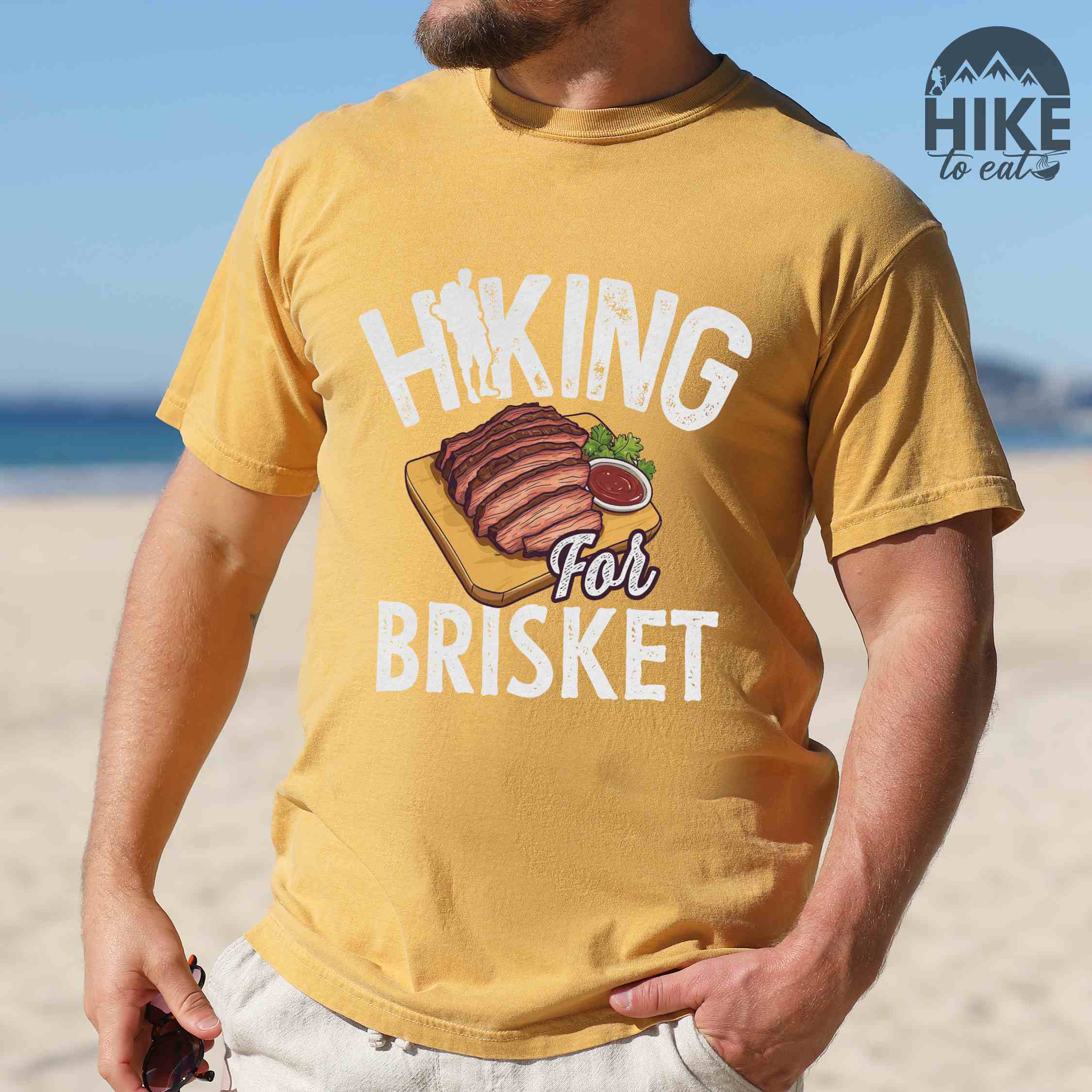 Man wearing mustard Hiking for brisket colored vintage tee with bold typography, illustrated sliced brisket, birds, and backpacker for brisket lovers