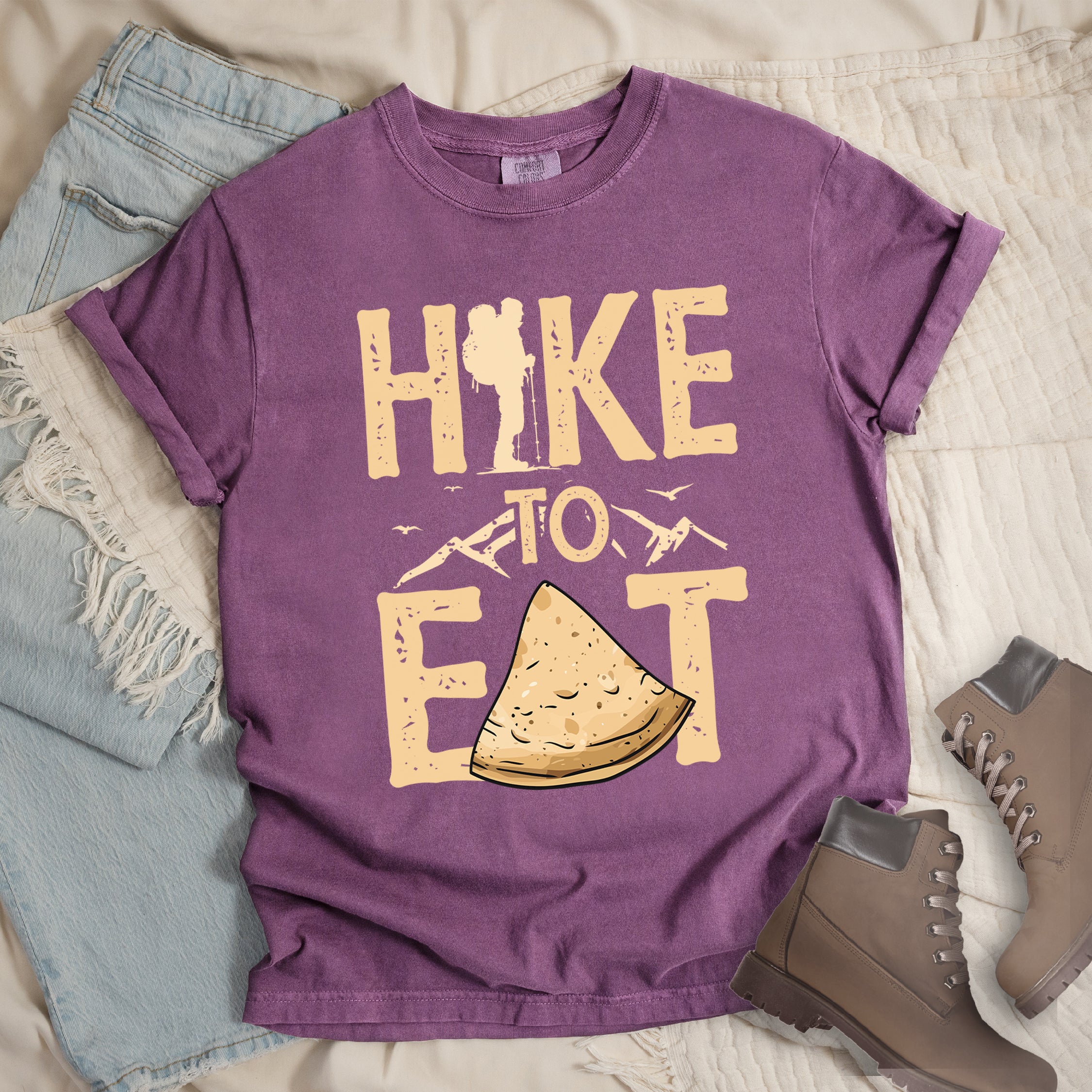 Berry (Dark Violet) t-shirt with text  "HIKE TO EAT". "HIKE" appears in large beige letters at the top with a hiker silhouette as letter "I", "TO" in beige text with small mountain outlines on either side in the middle, and "EAT" in large beige letters at the bottom with a triangular slice of flatbread or pastry (tortilla) replacing the letter "A". Small flying birds appear near "TO". Features a classic crew neck and casual rolled sleeves, styled alongside light wash denim and brown hiking boots. 