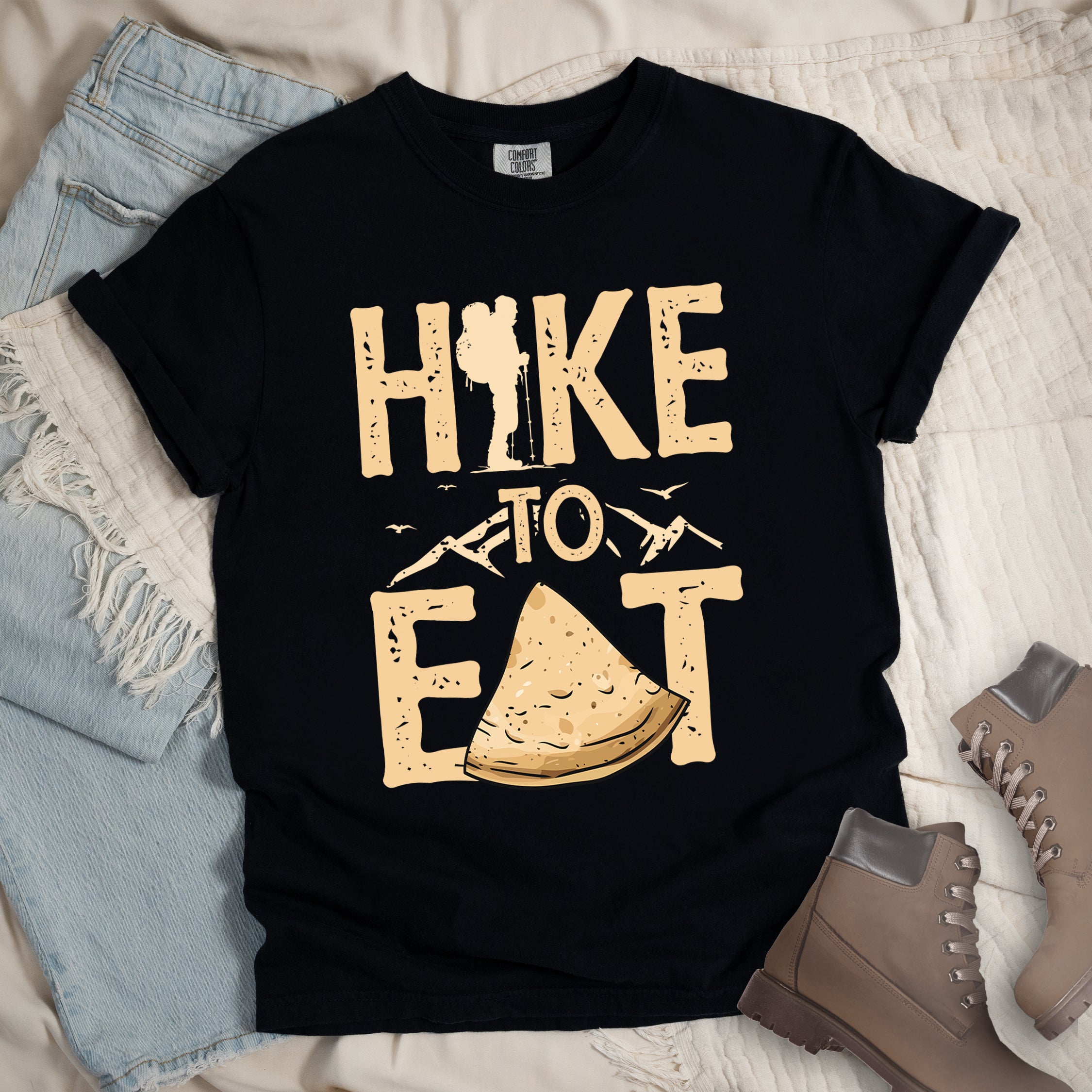 Black t-shirt with text  "HIKE TO EAT". "HIKE" appears in large beige letters at the top with a hiker silhouette as letter "I", "TO" in beige text with small mountain outlines on either side in the middle, and "EAT" in large beige letters at the bottom with a triangular slice of flatbread or pastry (tortilla) replacing the letter "A". Small flying birds appear near "TO". Features a classic crew neck and casual rolled sleeves, styled alongside light wash denim and brown hiking boots. 
