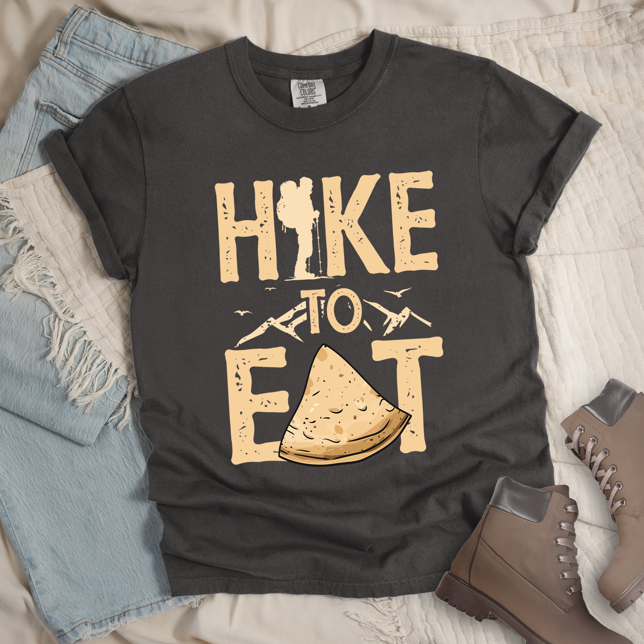 Pepper (Dark Gray) t-shirt with text  "HIKE TO EAT". "HIKE" appears in large beige letters at the top with a hiker silhouette as letter "I", "TO" in beige text with small mountain outlines on either side in the middle, and "EAT" in large beige letters at the bottom with a triangular slice of flatbread or pastry (tortilla) replacing the letter "A". Small flying birds appear near "TO". Features a classic crew neck and casual rolled sleeves, styled alongside light wash denim and brown hiking boots. 