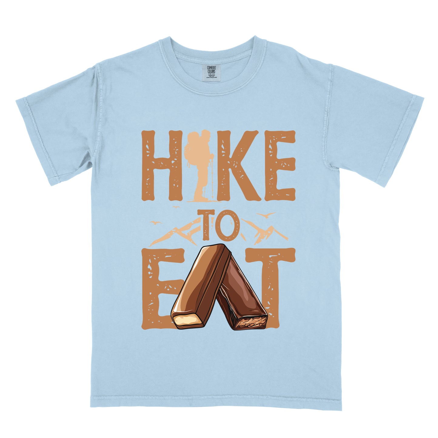Chambray (Light Blue) t-shirt with tan/brown distressed text reading "HIKE TO EAT". "HIKE" appears in large brown letters at the top with a hiker silhouette integrated into the letter "I", "TO" in brown text with small mountain outlines on either side in the middle, and "EAT" in large brown letters at the bottom with two chocolate candy bars arranged in an "A" shape replacing the letter "A". Small flying birds appear near "TO".
