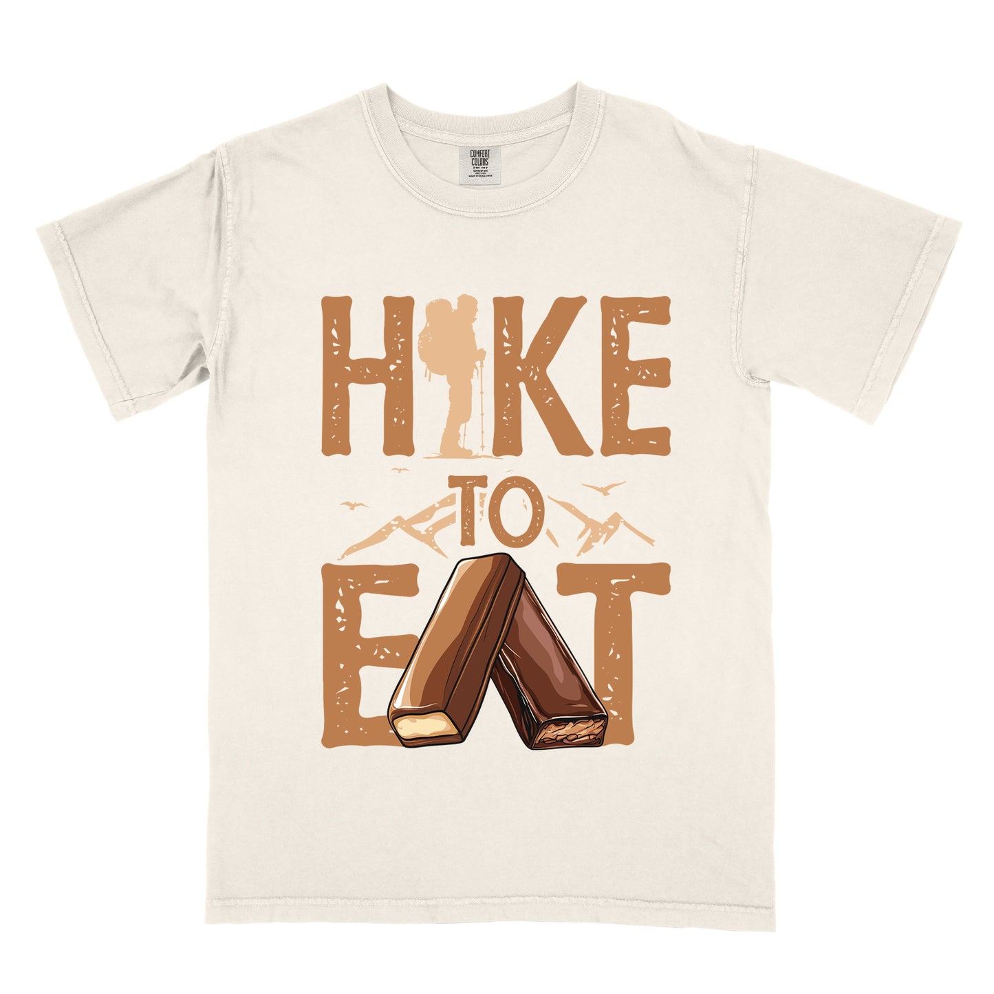 Ivory (Cream White) t-shirt with tan/brown distressed text reading "HIKE TO EAT". "HIKE" appears in large brown letters at the top with a hiker silhouette integrated into the letter "I", "TO" in brown text with small mountain outlines on either side in the middle, and "EAT" in large brown letters at the bottom with two chocolate candy bars arranged in an "A" shape replacing the letter "A". Small flying birds appear near "TO".