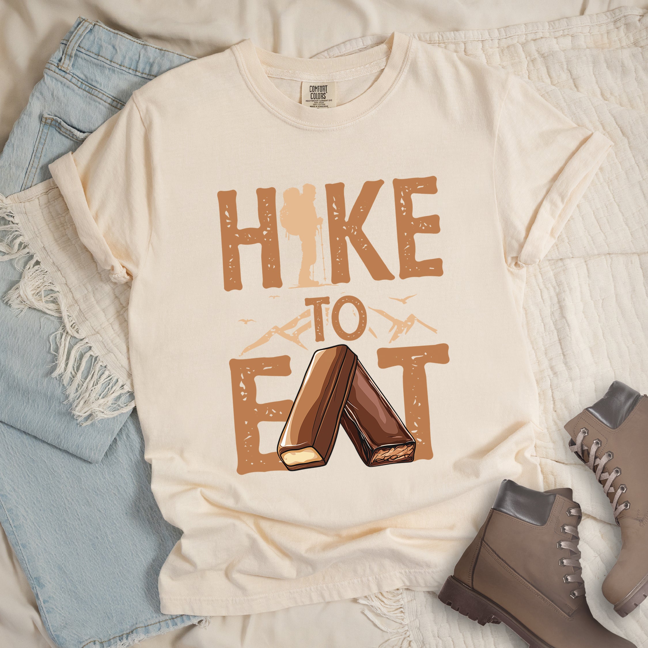 Ivory (Cream White) t-shirt with text "HIKE TO EAT". "HIKE" appears in large brown letters at the top with a hiker silhouette integrated into the letter "I", "TO" in brown text with small mountain outlines on either side in the middle, and "EAT" in large brown letters at the bottom with two chocolate candy bars arranged in an "A" shape replacing the letter "A". Small flying birds appear near "TO". Features a classic crew neck and casual rolled sleeves,  alongside light wash denim and brown hiking boots. 