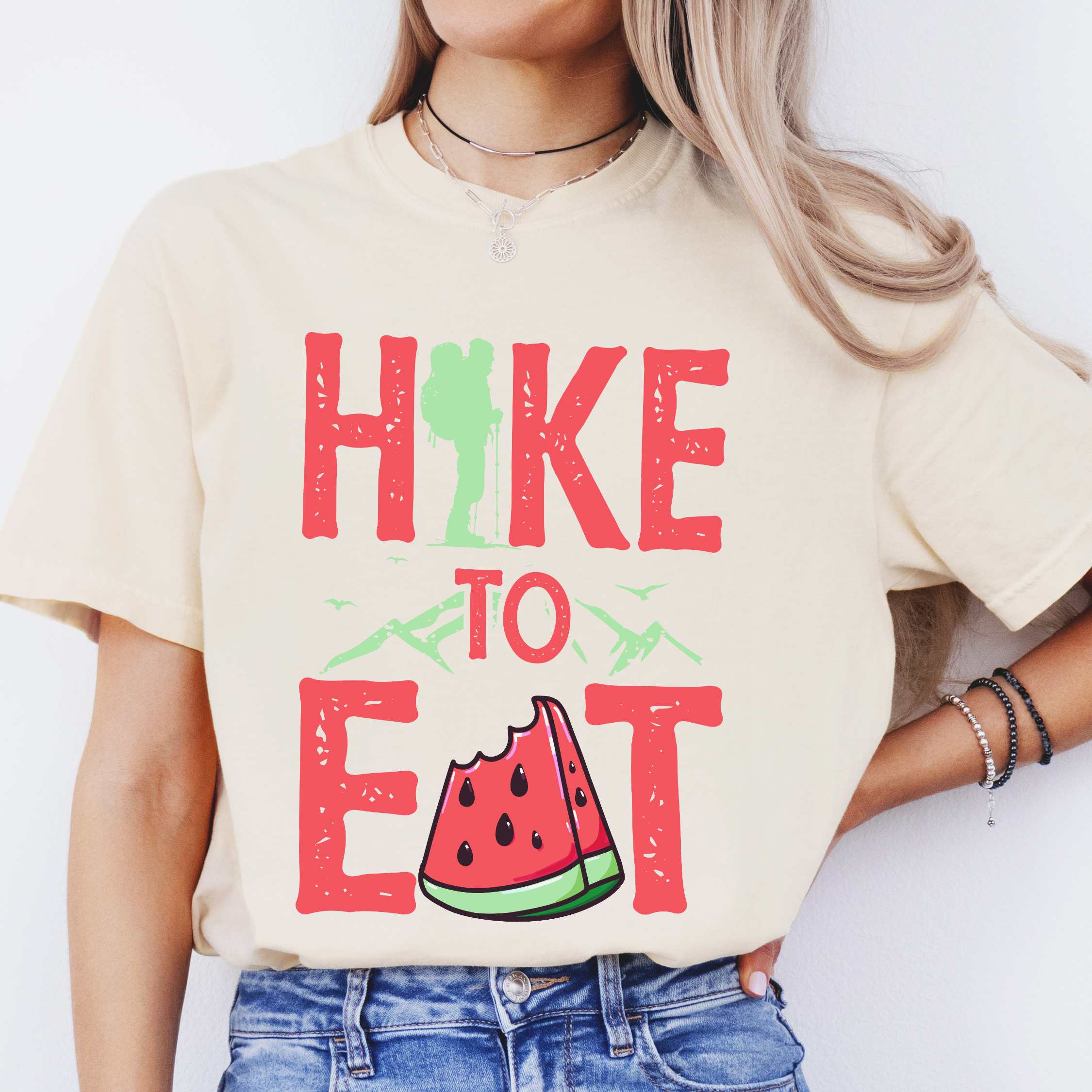Woman wearing ivory "Hike to Eat" colored statement tee with bold typography, mountain, illustrated slice of watermelon, perfect for fruit lovers.