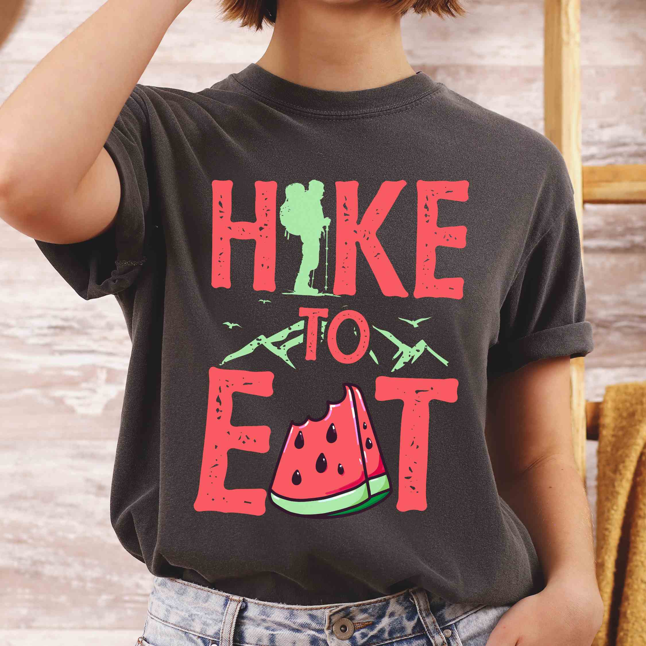Woman wearing pepper "Hike to Eat" colored statement tee with bold typography, mountain, illustrated slice of watermelon, perfect for fruit lovers.