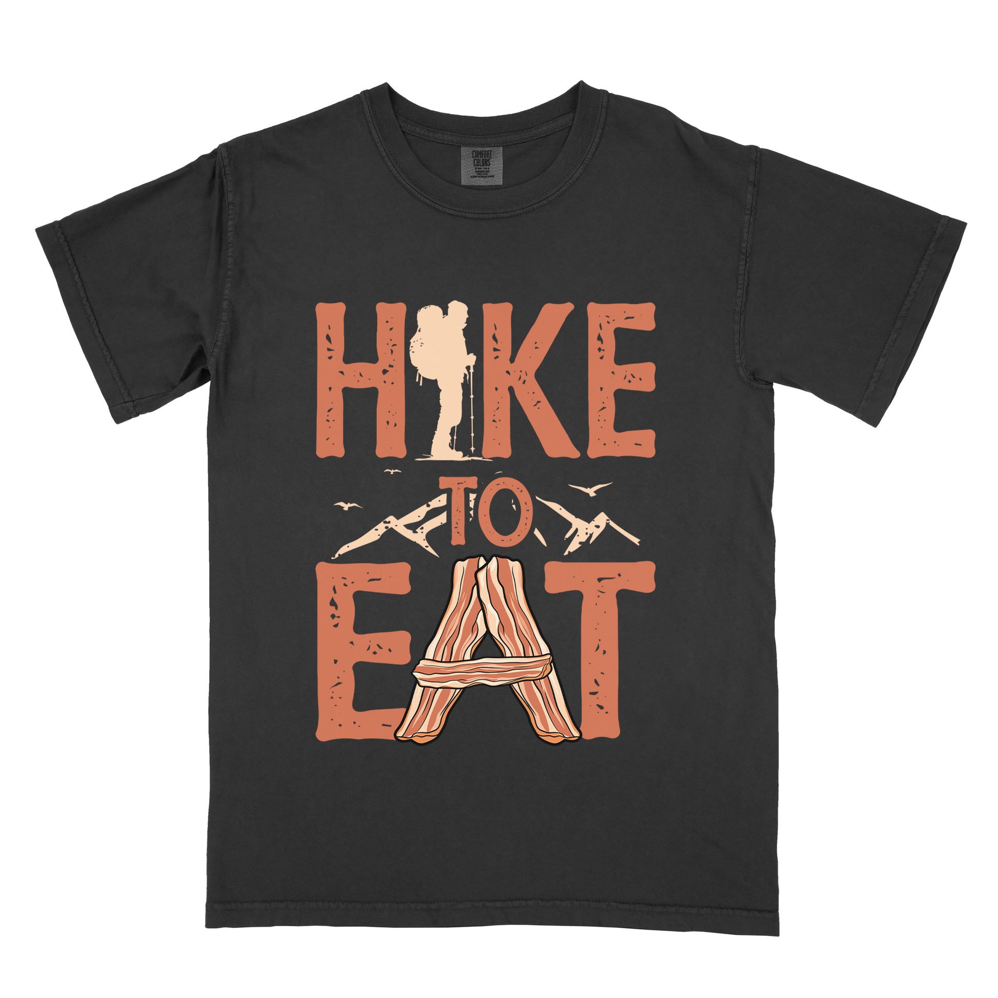 Black t-shirt with orange distressed text reading "HIKE TO EAT". "HIKE" appears in large orange letters at the top with a beige hiker silhouette integrated into the letter "I", "TO" in orange text with mountain outlines on either side in the middle, and "EAT" in large orange letters at the bottom with strips of bacon arranged in an "A" shape replacing the letter "A". Small flying birds appear near "TO". 