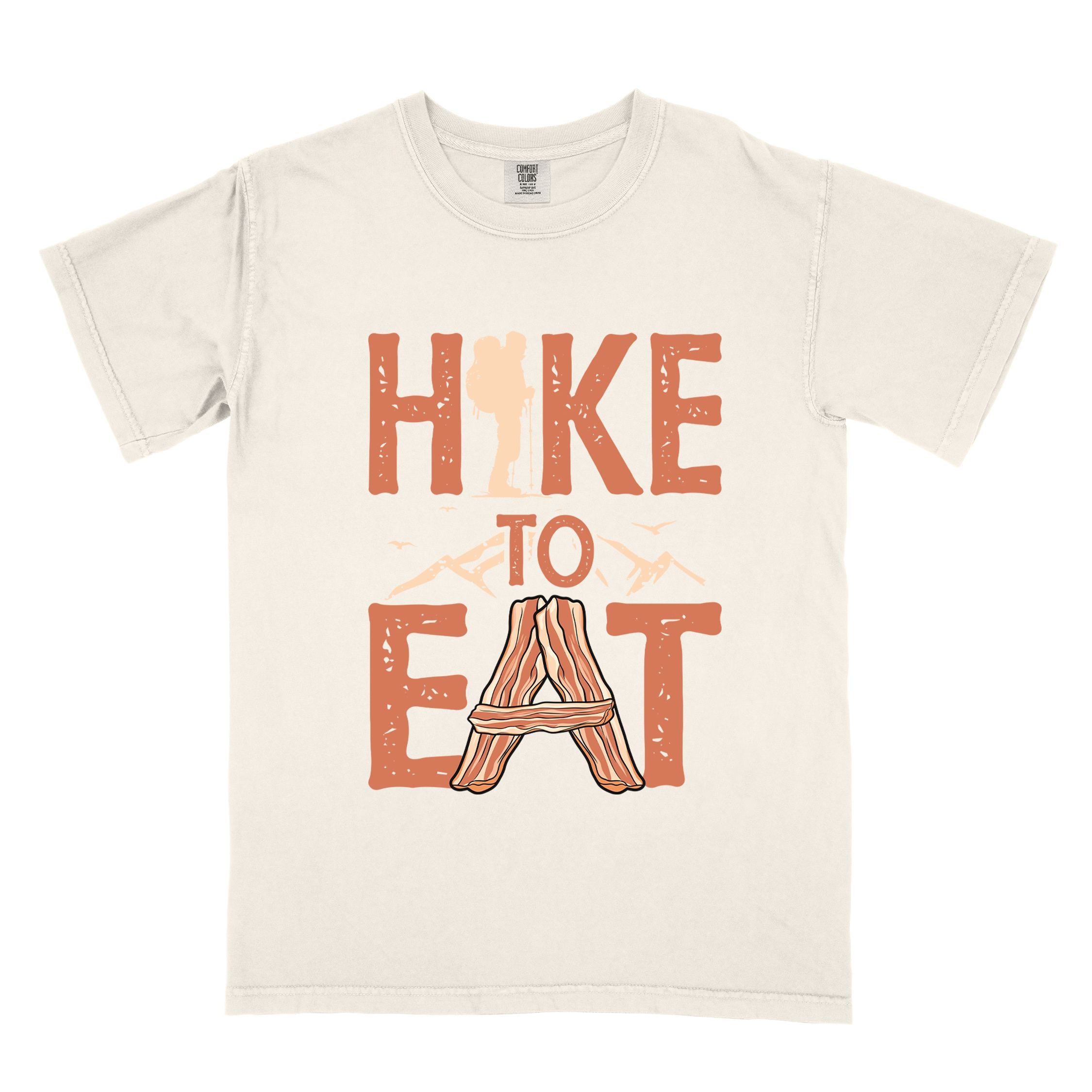Ivory "Hike to Eat" colored statement tee with bold typography, mountain, illustrated slices of bacon, and backpacker for breakfast enthusiasts.
