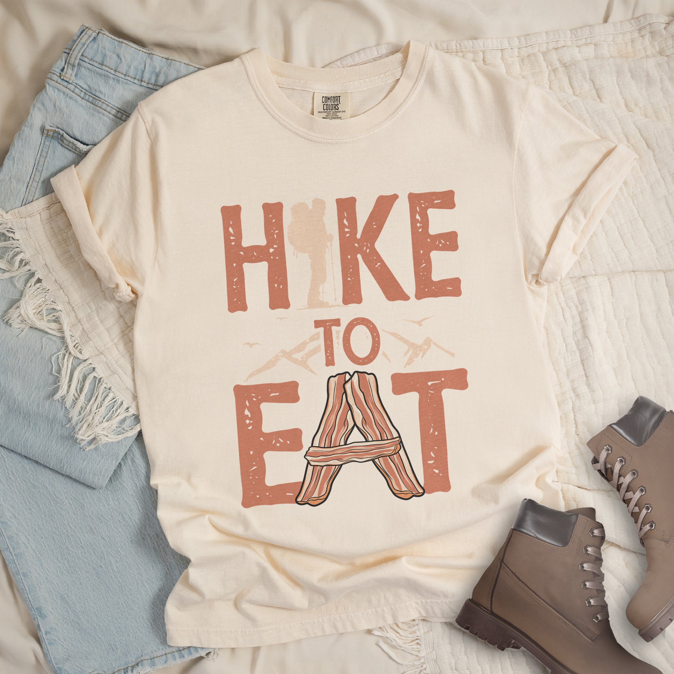 Ivory (Cream White) t-shirt with text reading "HIKE TO EAT". "HIKE" appears in large orange letters at the top with a beige hiker silhouette integrated into the letter "I", "TO" in orange text with mountain outlines on either side in the middle, and "EAT" in large orange letters at the bottom with strips of bacon arranged in an "A" shape replacing the letter "A". Small flying birds appear near "TO".  Features a classic crew neck and casual rolled sleeves, styled alongside light wash denim and brown boots. 