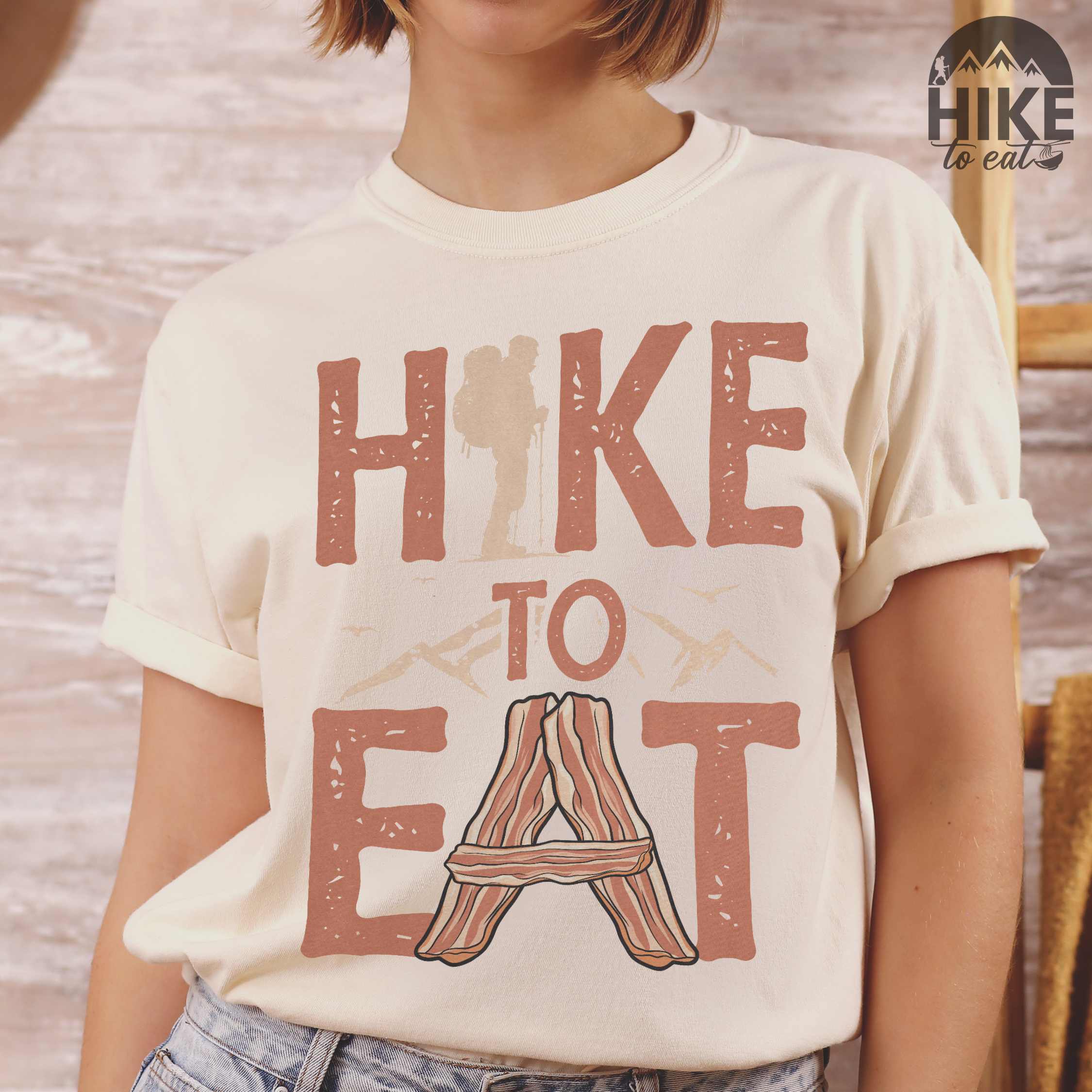woman wearing ivory "Hike to Eat" colored statement tee with bold typography, mountain, illustrated slices of bacon, and backpacker for breakfast enthusiasts.