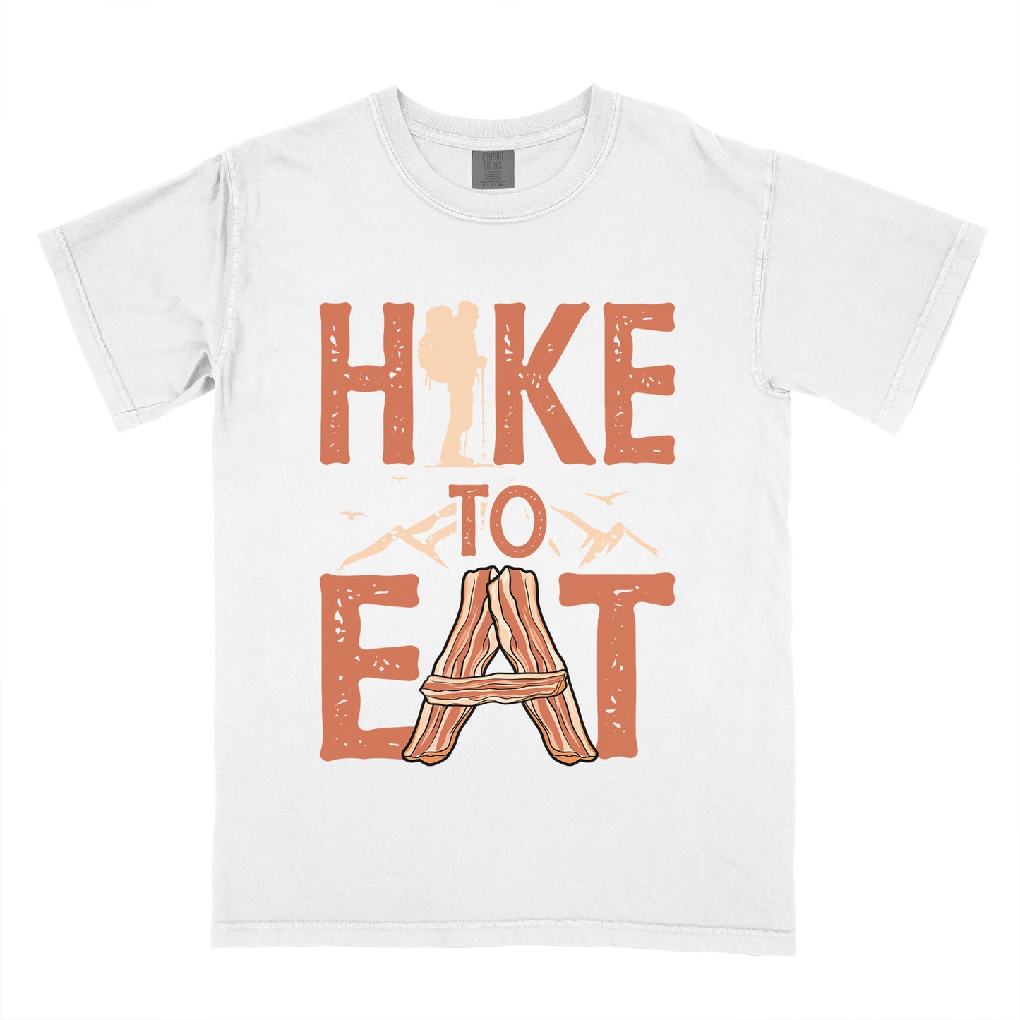White t-shirt with orange distressed text reading "HIKE TO EAT". "HIKE" appears in large orange letters at the top with a beige hiker silhouette integrated into the letter "I", "TO" in orange text with mountain outlines on either side in the middle, and "EAT" in large orange letters at the bottom with strips of bacon arranged in an "A" shape replacing the letter "A". Small flying birds appear near "TO". 