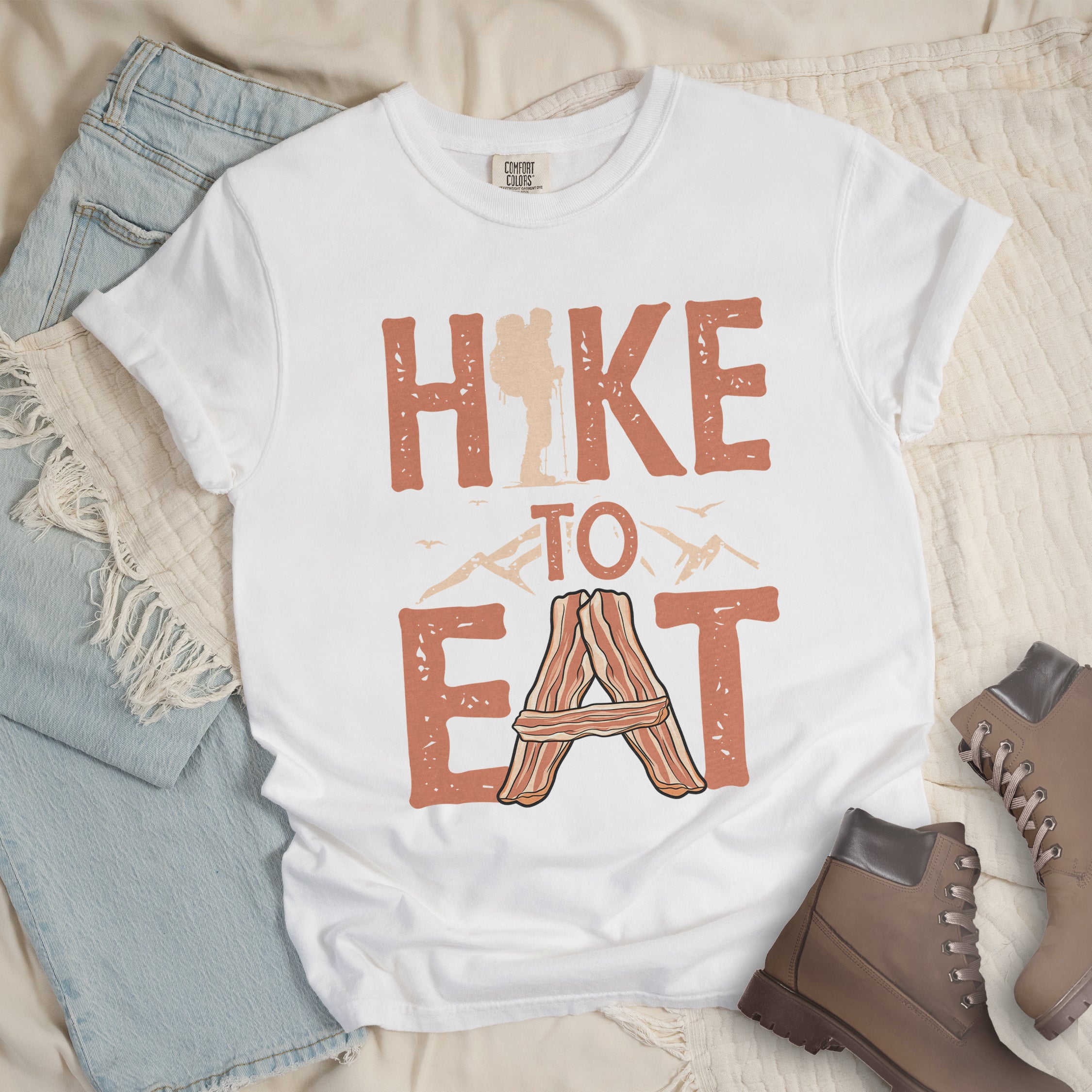 White t-shirt with text reading "HIKE TO EAT". "HIKE" appears in large orange letters at the top with a beige hiker silhouette integrated into the letter "I", "TO" in orange text with mountain outlines on either side in the middle, and "EAT" in large orange letters at the bottom with strips of bacon arranged in an "A" shape replacing the letter "A". Small flying birds appear near "TO".  The shirt features a classic crew neck and casual rolled sleeves, styled alongside light wash denim and brown hiking boots