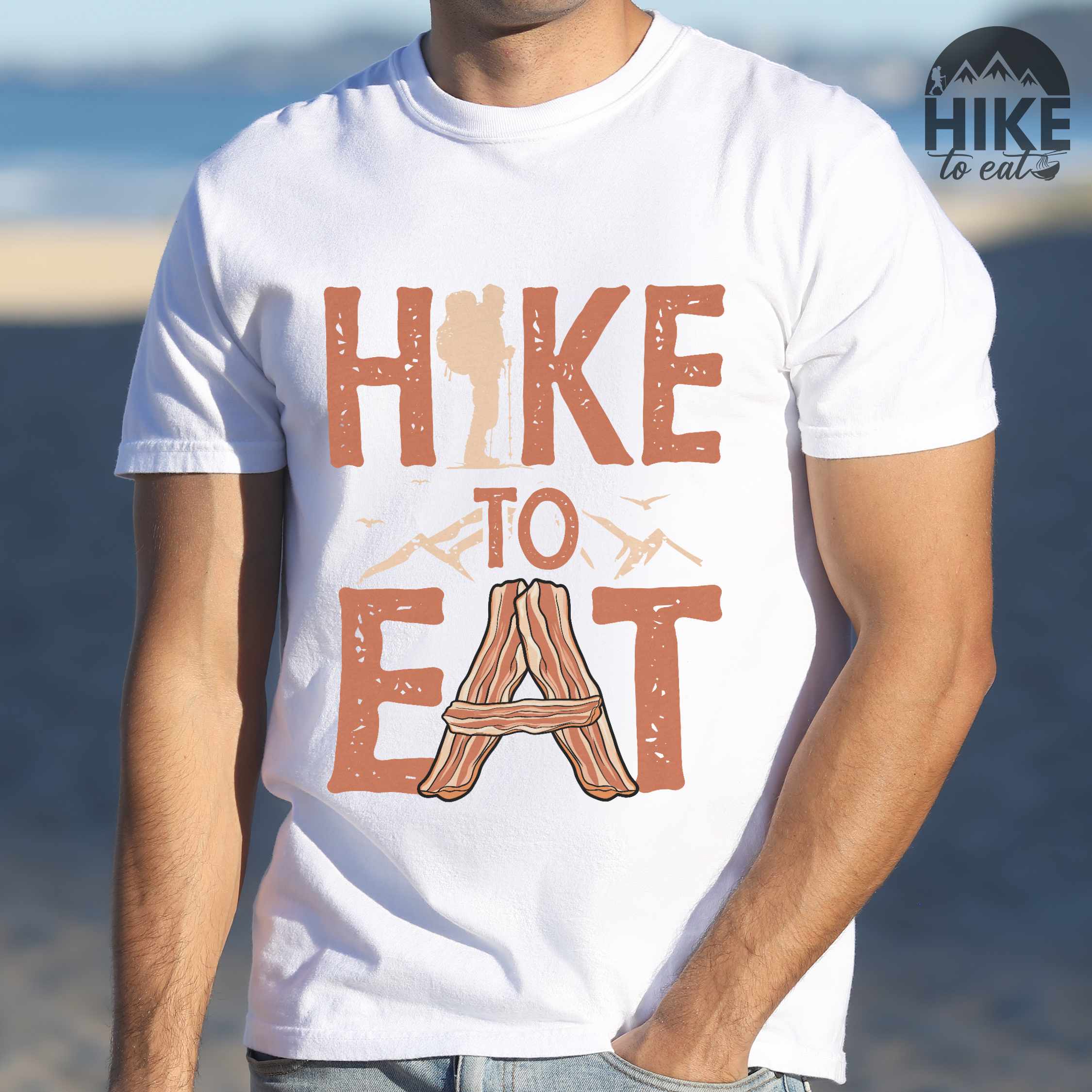 Man wearing white"Hike to Eat" colored statement tee with bold typography, mountain, illustrated slices of bacon, and backpacker for breakfast enthusiasts.