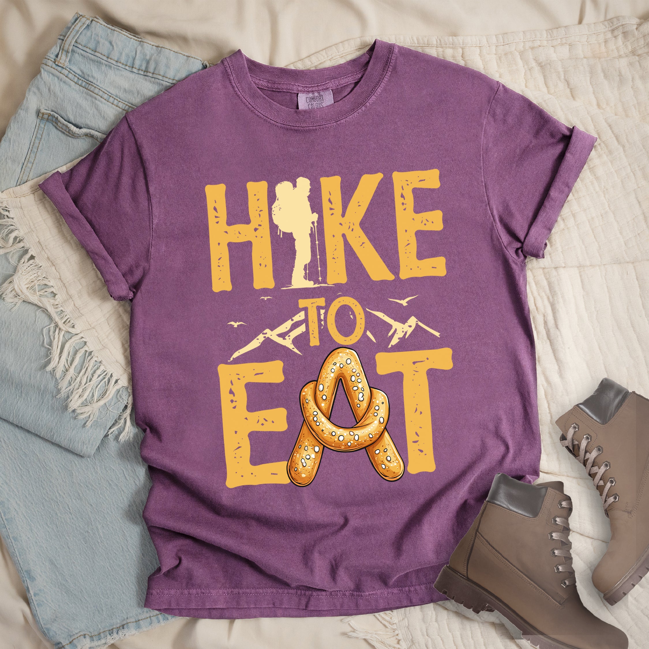 Berry (Dark Violet) t-shirt with text reading "HIKE TO EAT". "HIKE" appears in golden yellow letters at the top with a hiker silhouette integrated into the letter "I", "TO" in golden yellow text with mountain outlines on either side in the middle, and "EAT" in large golden yellow letters at the bottom with a pretzel shaped in an "A" replacing the letter "A". Small flying birds appear near "TO".  Features a classic crew neck and casual rolled sleeves, styled alongside light wash denim and brown hiking boots.