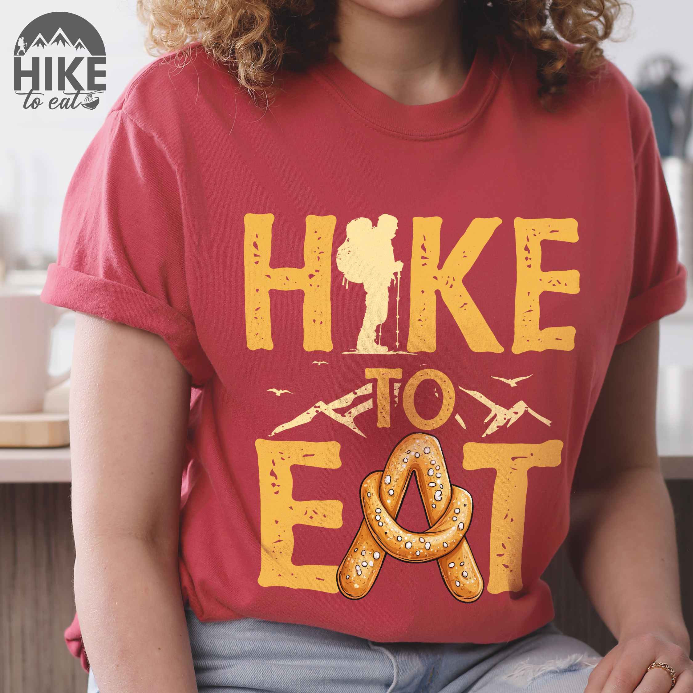 Woman wearing crimson "Hike to Eat" colored statement tee with bold typography, mountain, illustrated baked pretzel, and backpacker for pastry enthusiasts.