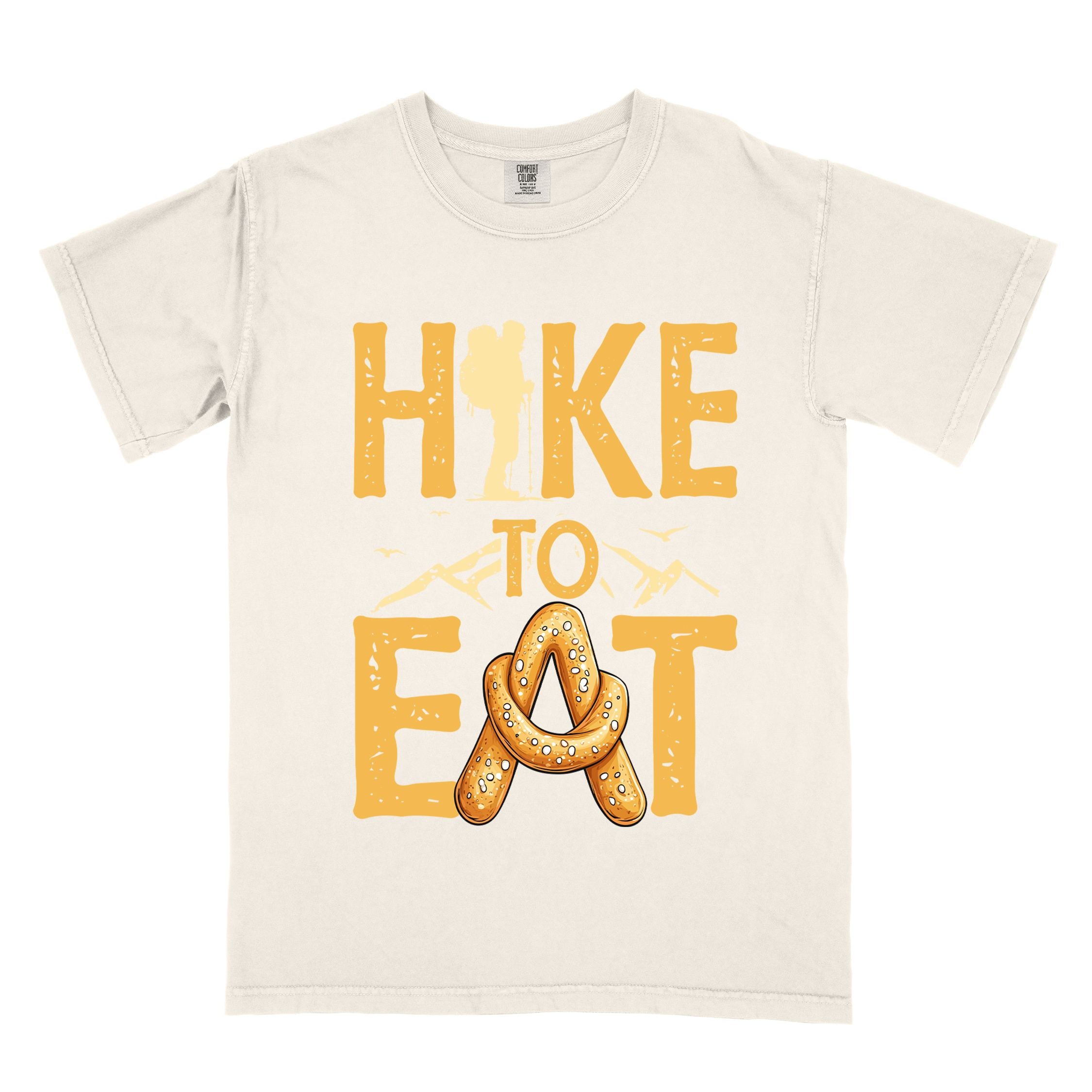 Ivory "Hike to Eat" colored statement tee with bold typography, mountain, illustrated baked pretzel, and backpacker for pastry enthusiasts.