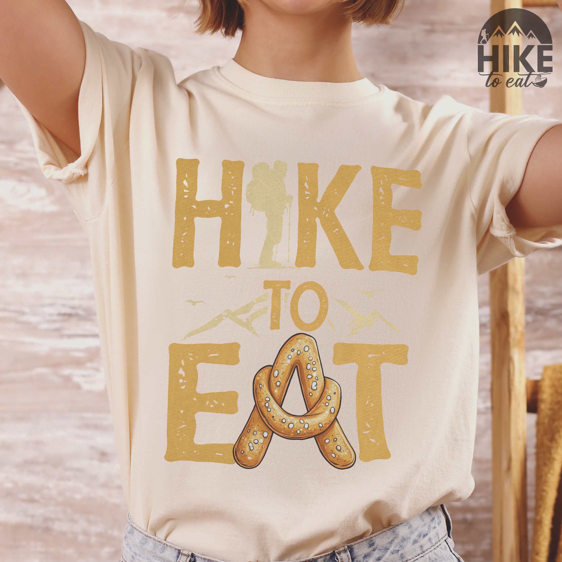 woman wearing ivory "Hike to Eat" colored statement tee with bold typography, mountain, illustrated baked pretzel, and backpacker for pastry enthusiasts.