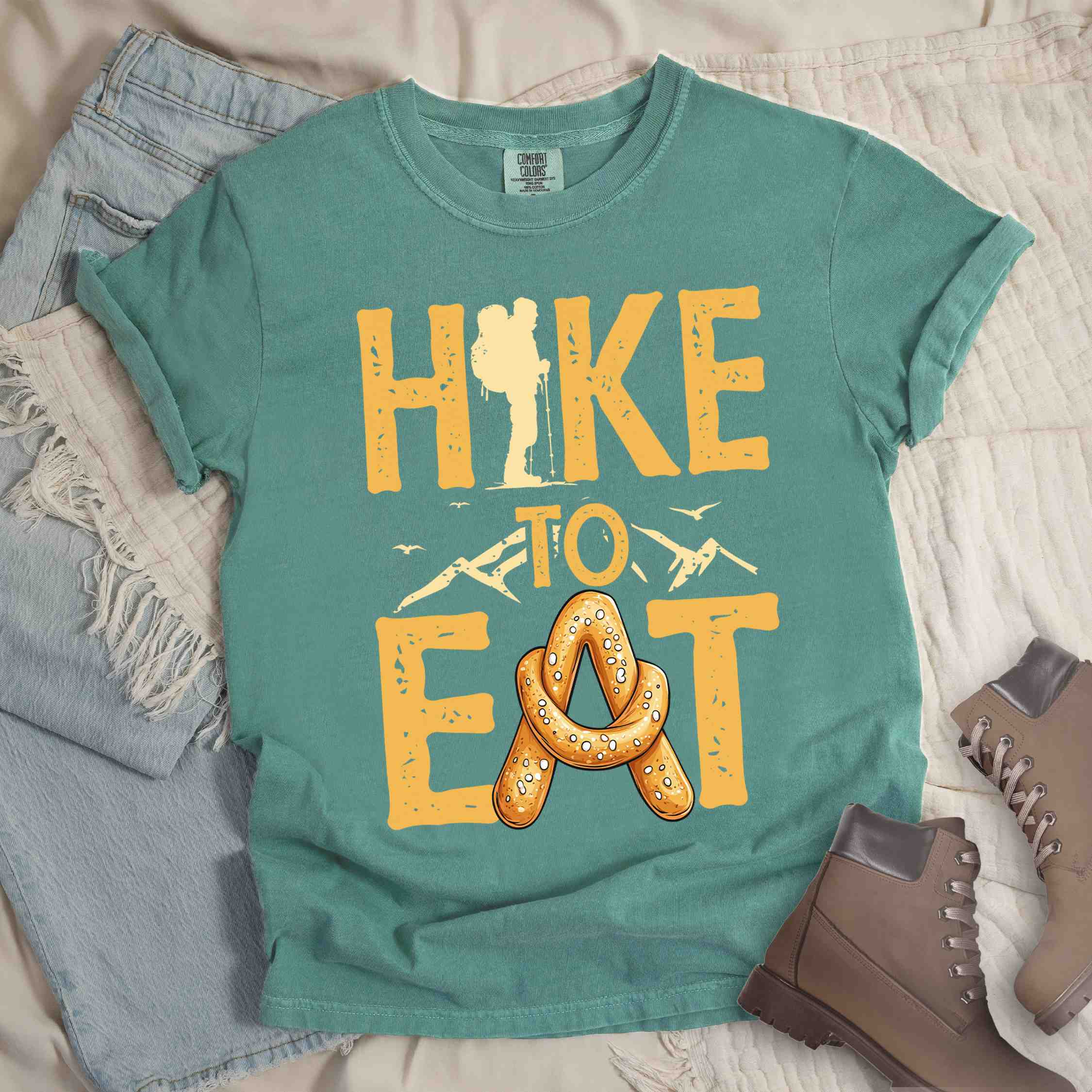 Light green "Hike to Eat" colored statement tee with bold typography, mountain, illustrated baked pretzel, and backpacker for pastry enthusiasts.