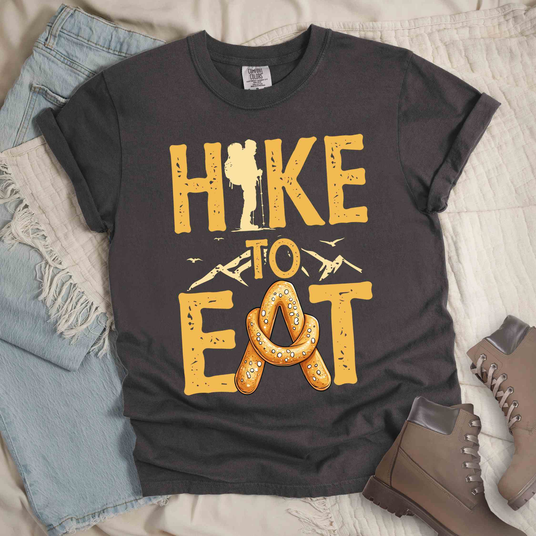 Pepper "Hike to Eat" colored statement tee with bold typography, mountain, illustrated baked pretzel, and backpacker for pastry enthusiasts.