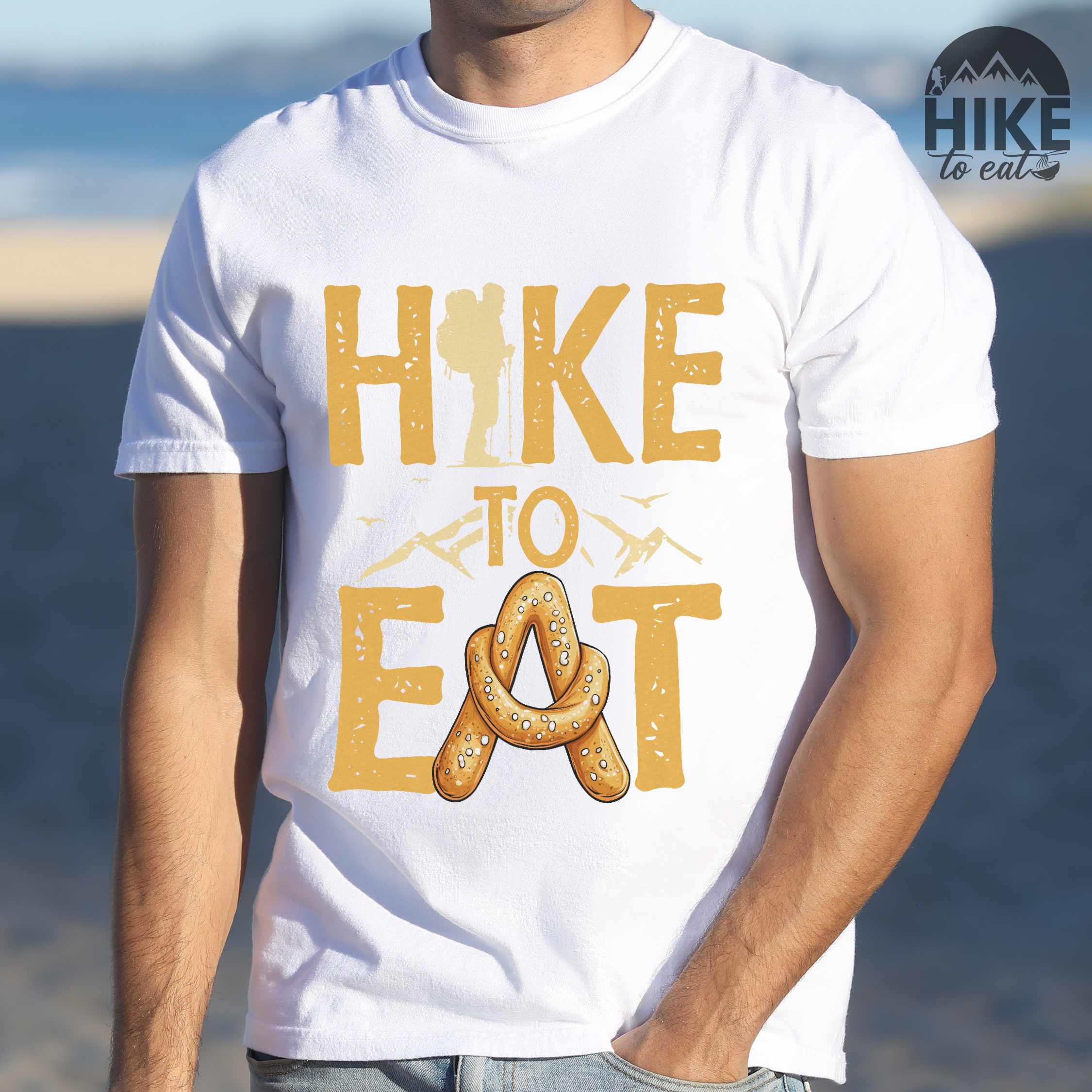 Man wearing white "Hike to Eat" colored statement tee with bold typography, mountain, illustrated baked pretzel, and backpacker for pastry enthusiasts.