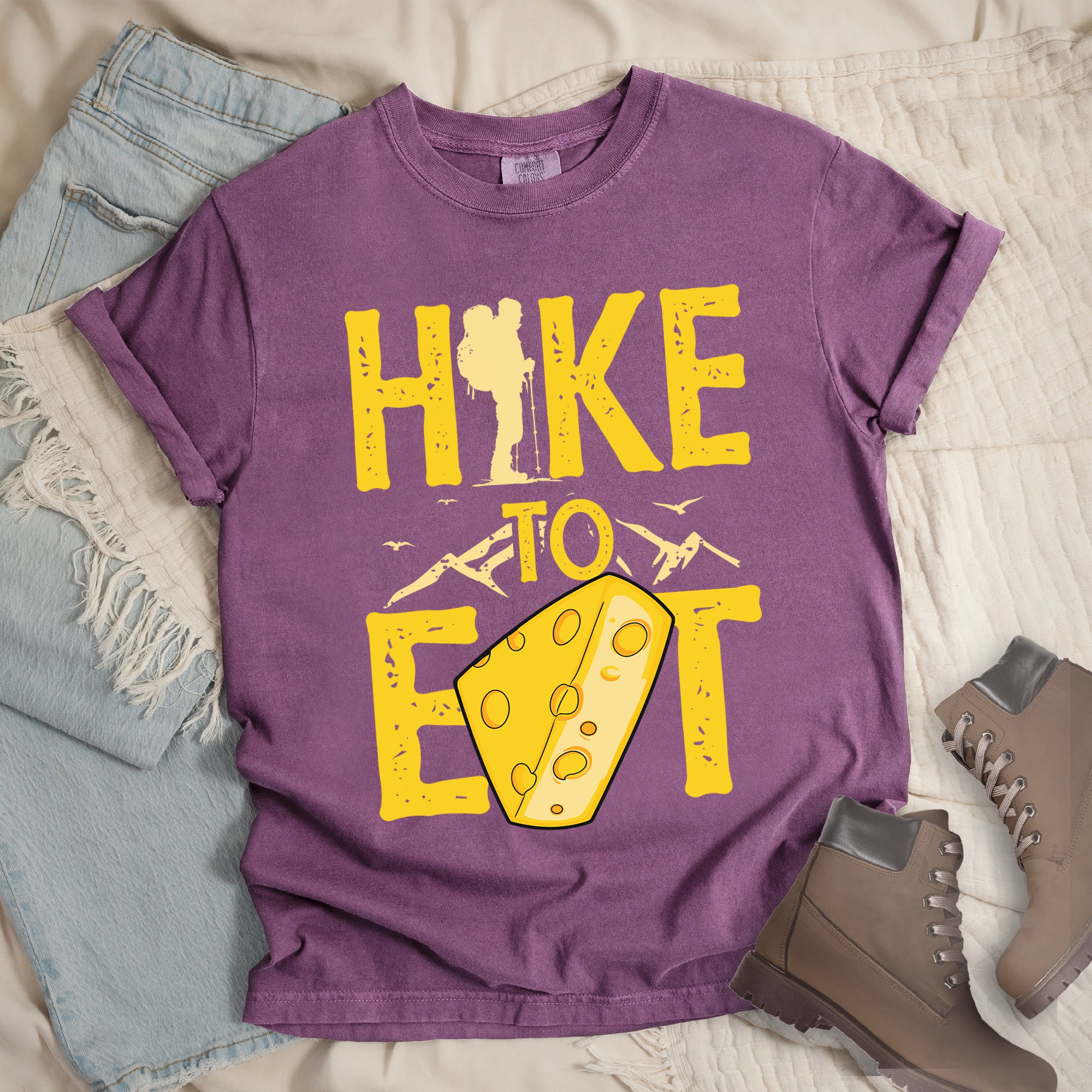 Berry (Dark Violet) t-shirt with yellow distressed text reading "HIKE TO EAT". "HIKE" appears in large yellow letters at the top with a hiker silhouette integrated into the letter "I", "TO" in yellow text with mountain outlines on either side in the middle, and "EAT" in large yellow letters at the bottom with a wedge of Swiss cheese positioned between "E" and "T". Small flying birds appear near "TO".  Features a classic crew neck and casual rolled sleeves, alongside light wash denim and brown hiking boots.
