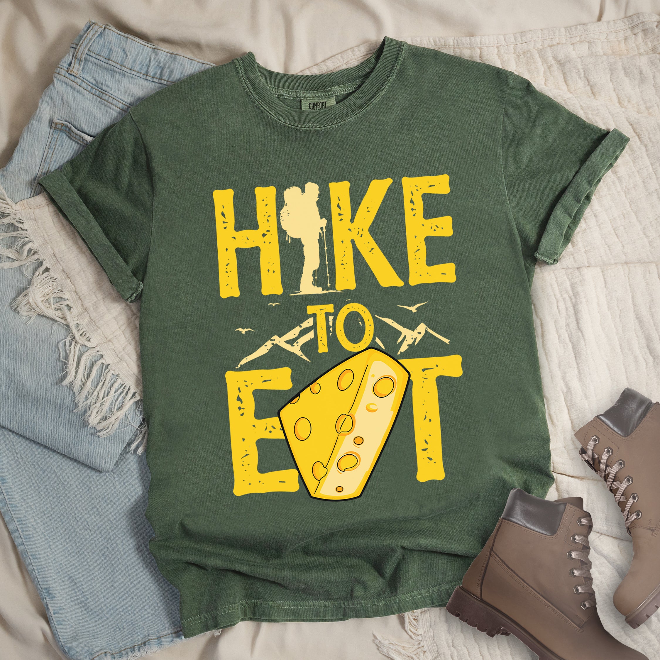 Moss (Dark Green) t-shirt with yellow distressed text reading "HIKE TO EAT". "HIKE" appears in large yellow letters at the top with a hiker silhouette integrated into the letter "I", "TO" in yellow text with mountain outlines on either side in the middle, and "EAT" in large yellow letters at the bottom with a wedge of Swiss cheese positioned between "E" and "T". Small flying birds appear near "TO". Features a classic crew neck and casual rolled sleeves, alongside light wash denim and brown hiking boots. 