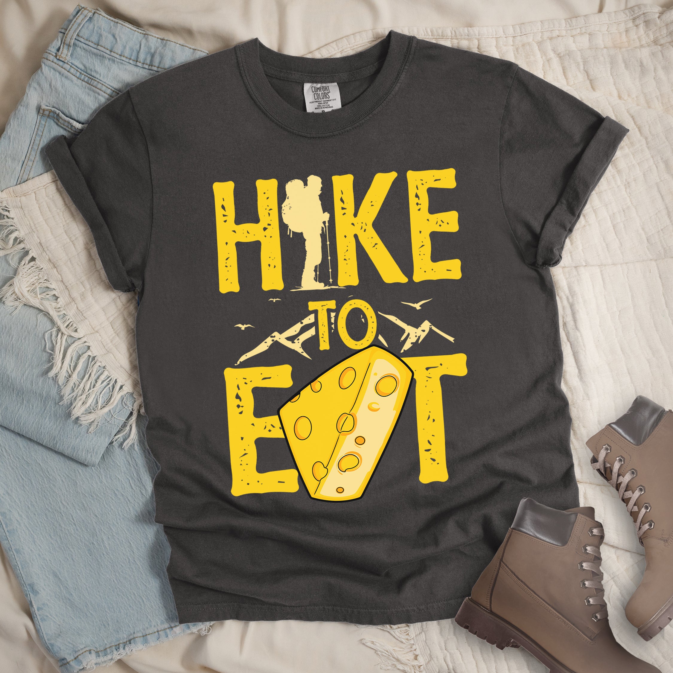 Pepper (Dark Gray) t-shirt with yellow distressed text reading "HIKE TO EAT". "HIKE" appears in large yellow letters at the top with a hiker silhouette integrated into the letter "I", "TO" in yellow text with mountain outlines on either side in the middle, and "EAT" in large yellow letters at the bottom with a wedge of Swiss cheese positioned between "E" and "T". Small flying birds appear near "TO".  Features a classic crew neck and casual rolled sleeves, alongside light wash denim and brown hiking boots.