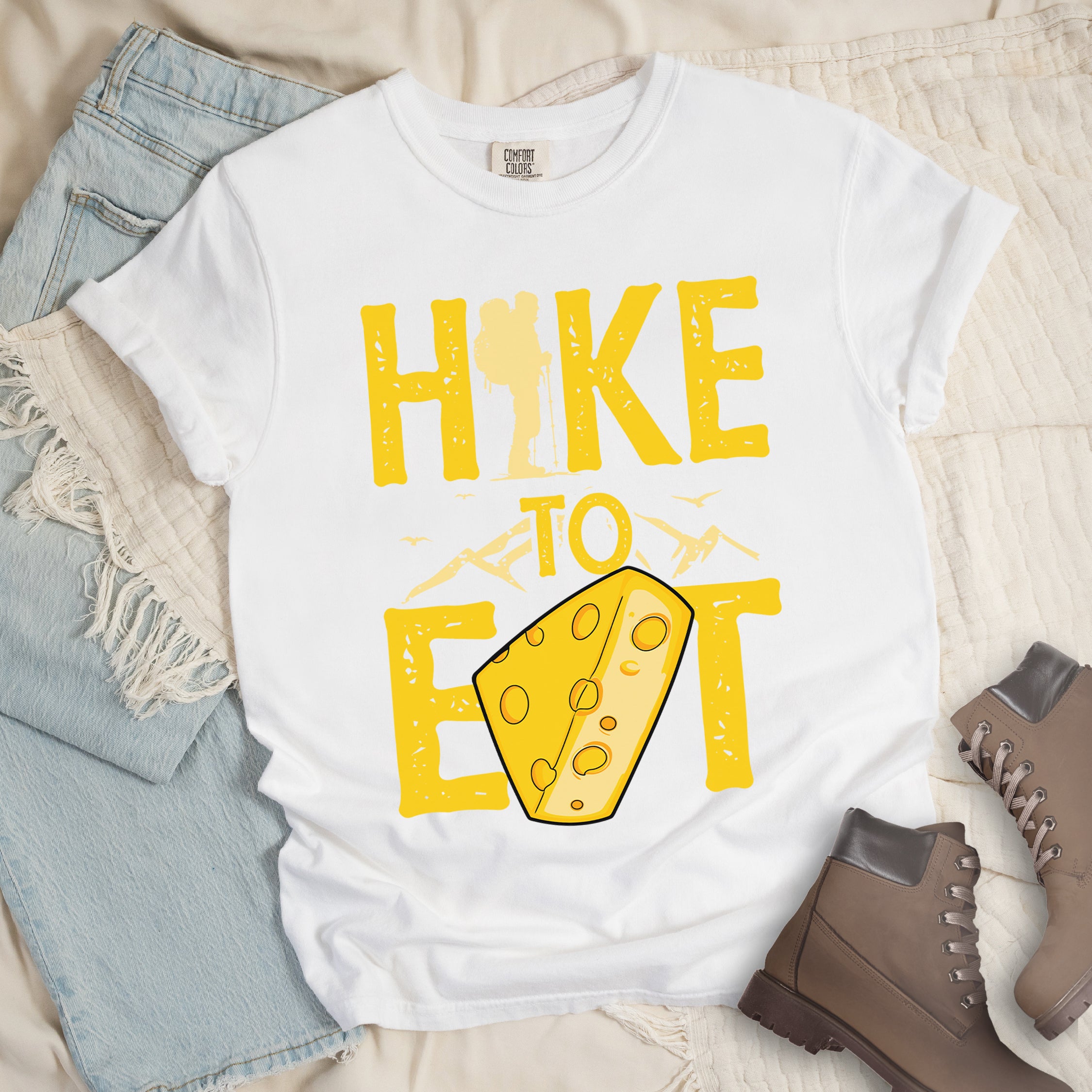 White t-shirt with yellow distressed text reading "HIKE TO EAT". "HIKE" appears in large yellow letters at the top with a hiker silhouette integrated into the letter "I", "TO" in yellow text with mountain outlines on either side in the middle, and "EAT" in large yellow letters at the bottom with a wedge of Swiss cheese positioned between "E" and "T". Small flying birds appear near "TO".  Features a classic crew neck and casual rolled sleeves, alongside light wash denim and brown hiking boots.