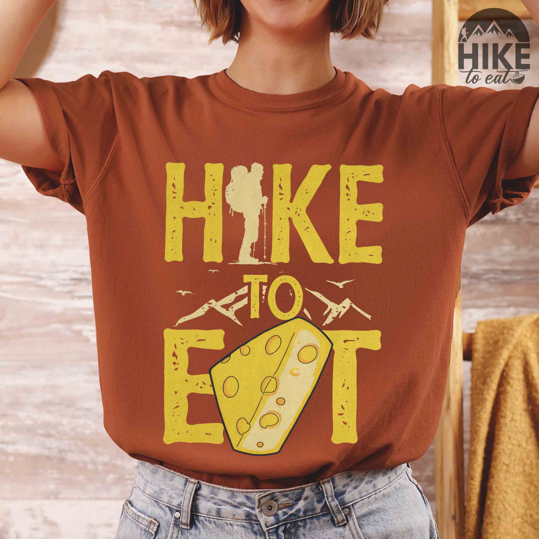 Woman wearing yam "Hike to Eat" colored statement tee with bold typography, mountain, illustrated slice of cheese, and backpacker for cheese lovers.