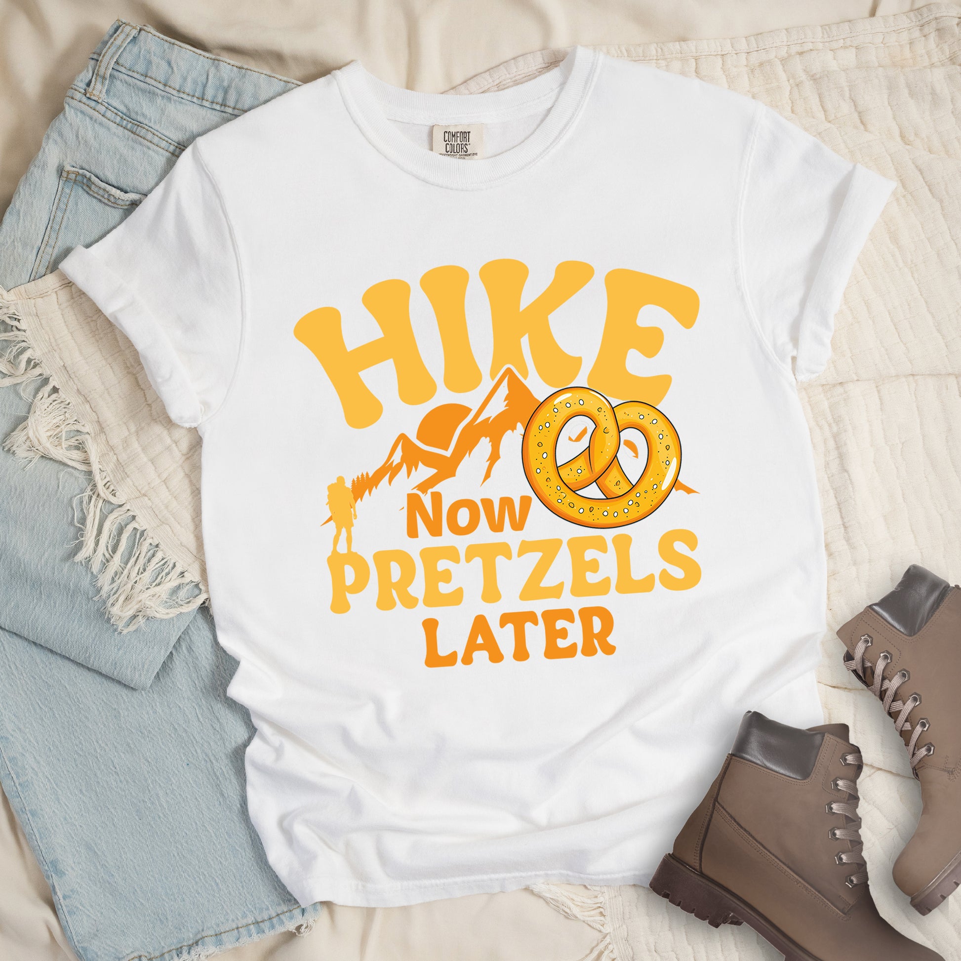 White t-shirt with "HIKE NOW PRETZELS LATER" text design. "HIKE" appears at the top in yellow letters, "NOW" in orange, "PRETZELS" in yellow, and "LATER" in orange. The design includes orange mountain silhouettes with a small yellow hiker figure and an illustration of a golden pretzel with salt. The playful text arrangement emphasizes hiking first and enjoying pretzels as a reward afterward. Features a classic crew neck and casual rolled sleeves, styled alongside light wash denim and brown hiking boots. 