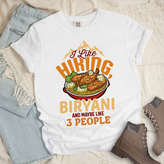 White t-shirt with text reading "I Like HIKING, BIRYANI AND MAYBE LIKE 3 PEOPLE". Gold mountain silhouettes appear above "I Like" (in dark brown script). "HIKING" is in large orange letters, below which sits a detailed illustration of a red bowl filled with biryani rice and chicken. "BIRYANI" appears in gold letters, while "AND MAYBE LIKE" is in small dark brown text and "3 PEOPLE" in orange. Features a classic crew neck and casual rolled sleeves, styled alongside light wash denim and brown hiking boots. 
