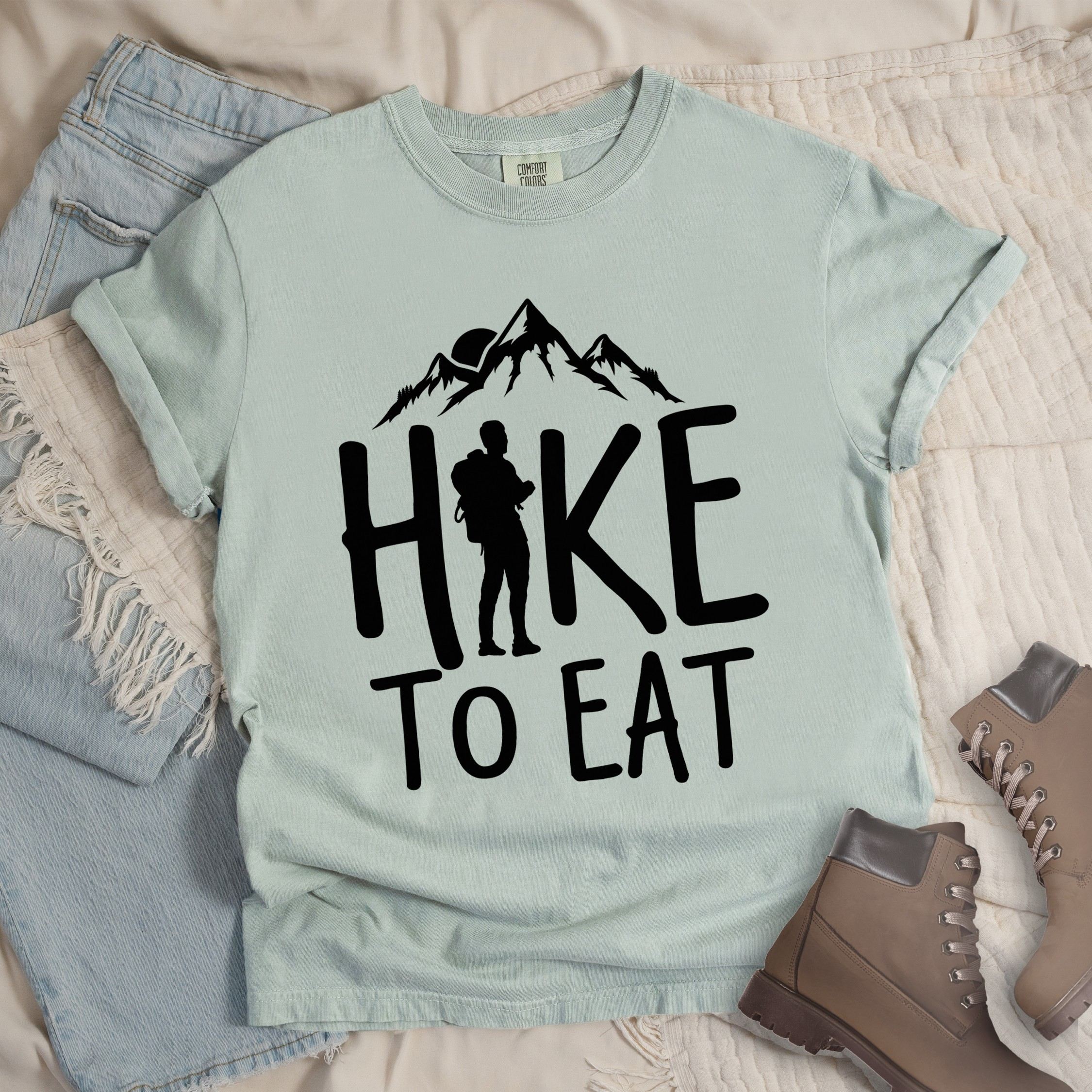 Light grey shirt with a Hiking design featuring mountain silhouette and shows 'Hike To Eat' phrase in bold text
