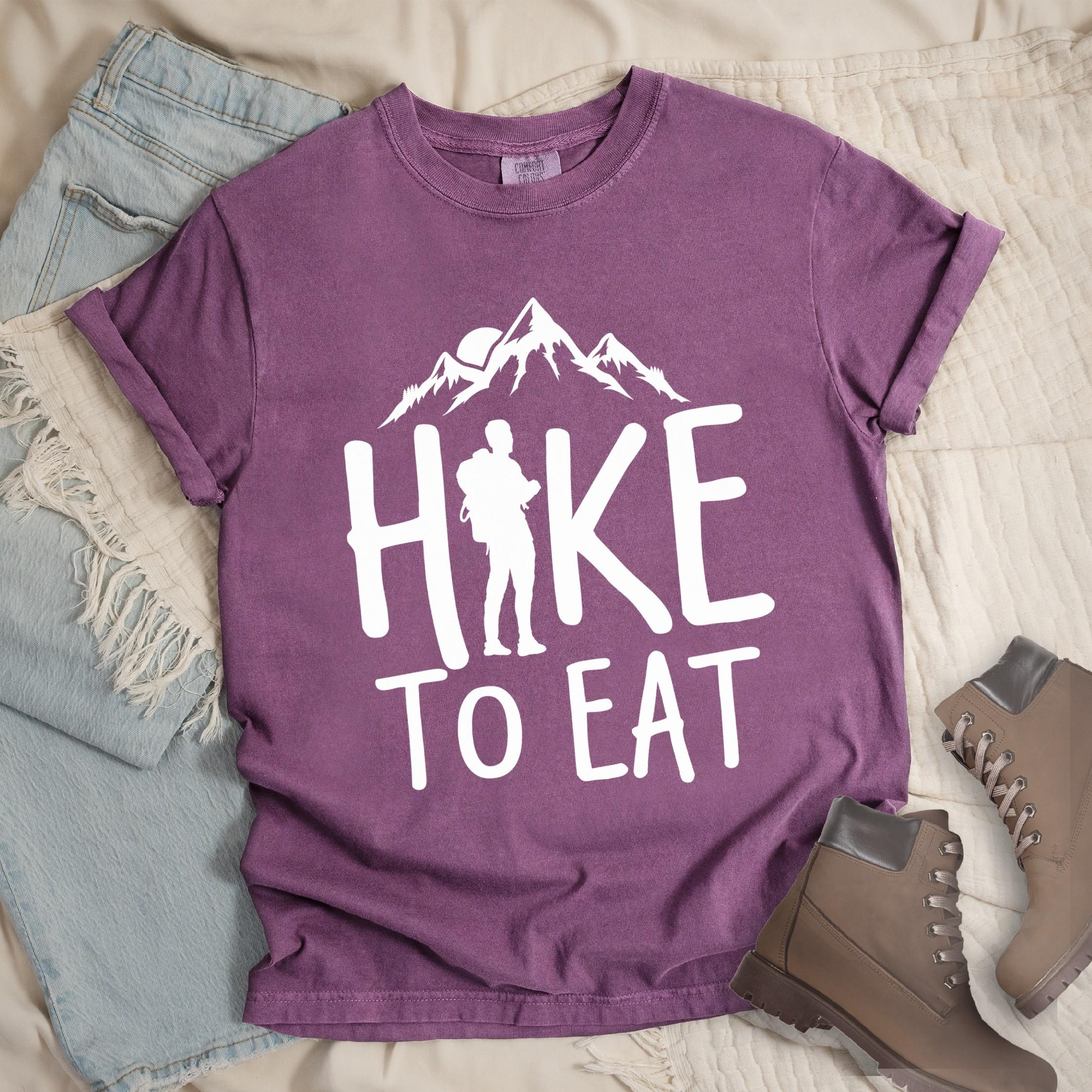 Dark pink shirt with a Hiking design featuring mountain silhouette and shows 'Hike To Eat' phrase in bold text