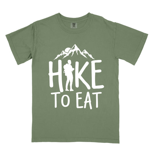 Moss (Dark Green) t-shirt with white text reading "HIKE TO EAT". Mountain silhouettes appear above the word "HIKE". The letter "I" in "HIKE" is replaced with a silhouette of a standing hiker with backpack. All text and graphics are in white against the Moss (Dark Green) shirt.